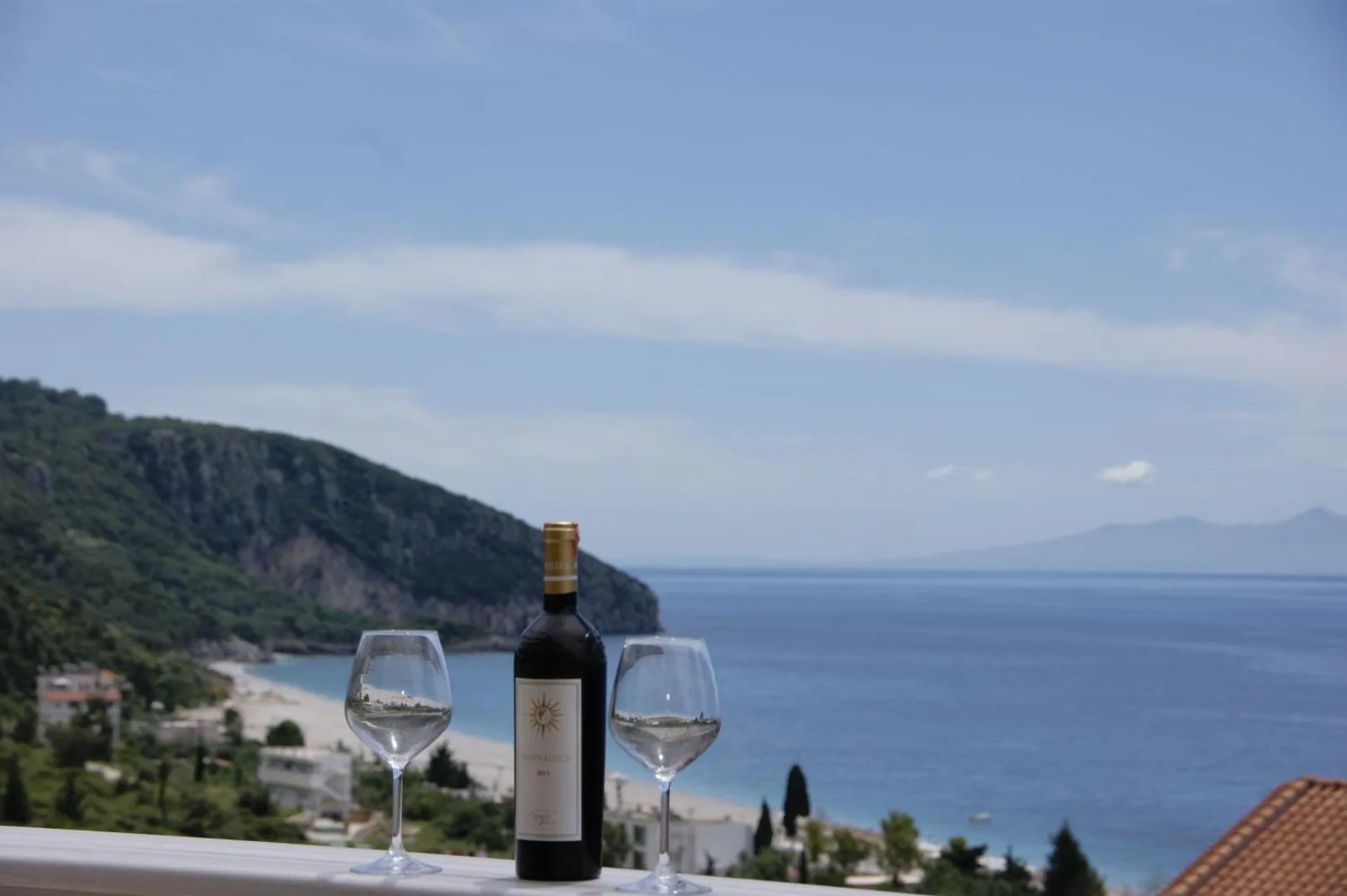 Two wine glasses and a bottle on a balcony rail, with the long pale curve of Dhërmi Beach, a wooded headland and the sea beyond