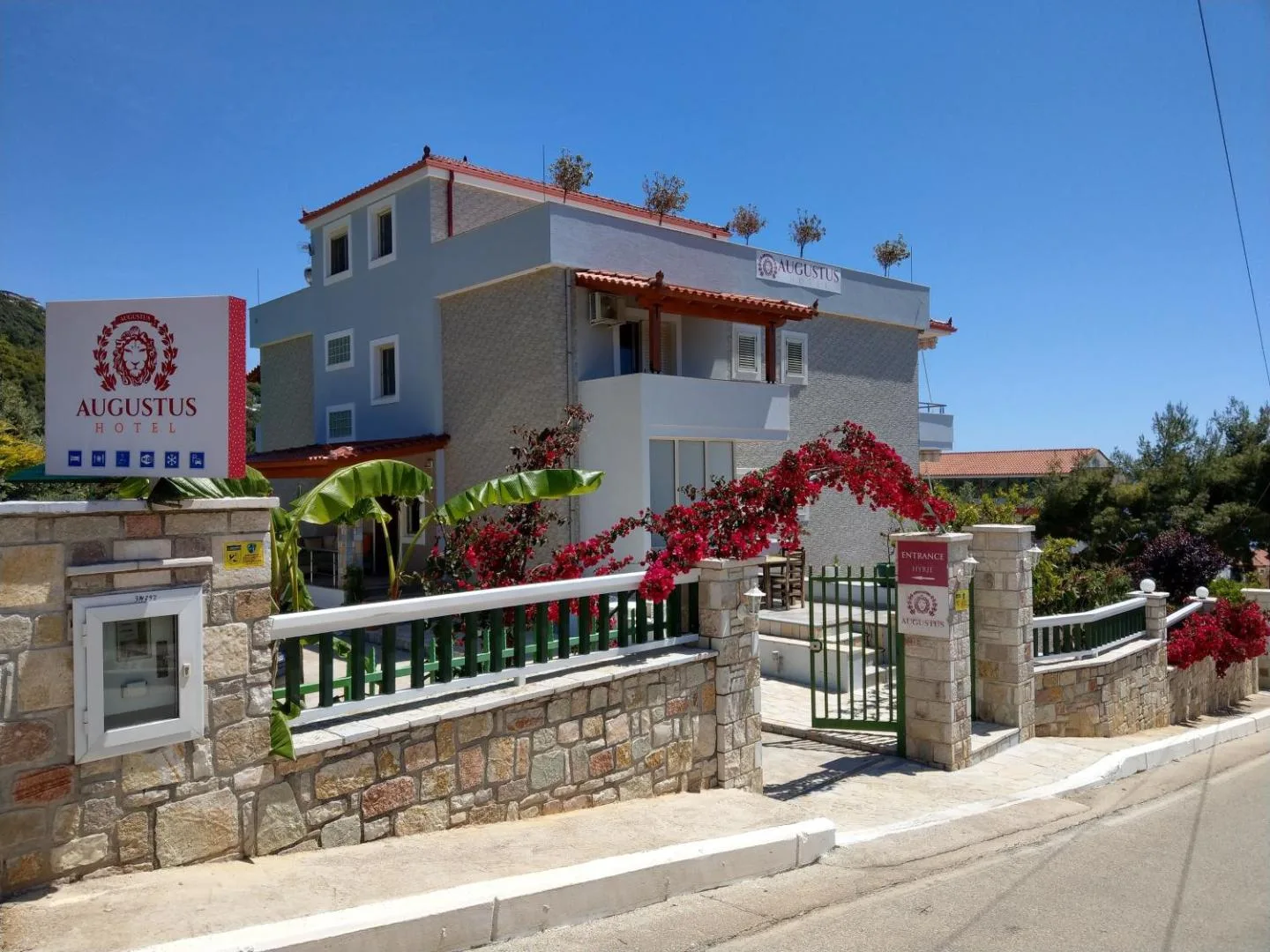 The front of Augustus Hotel from the road: a blue-grey three-storey building above a dressed-stone wall, a green gate under a red bougainvillea, and a white sign bearing a claret lion-in-laurel crest and the words Augustus Hotel