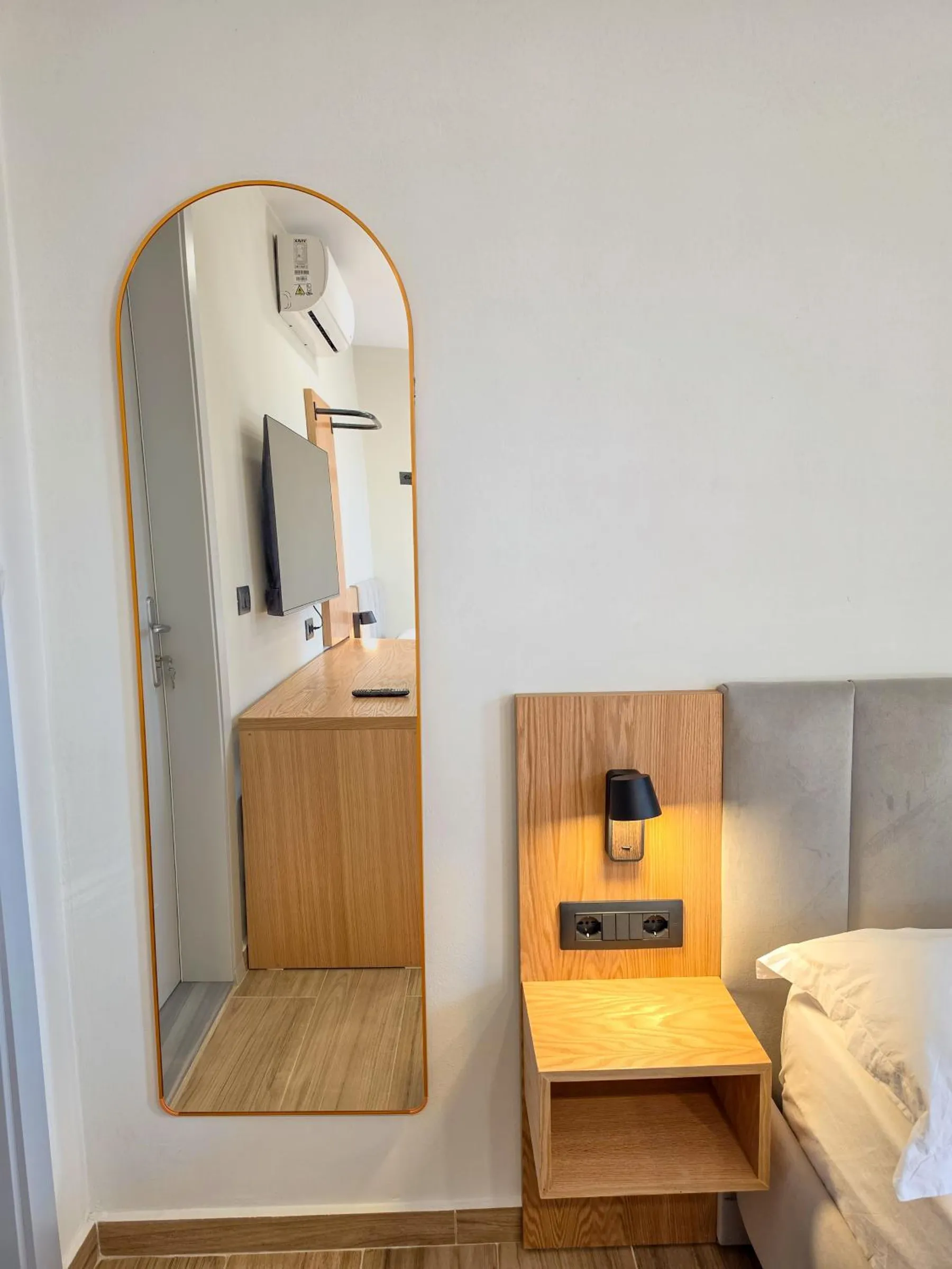 A tall arched mirror on a white wall beside an oak bedside shelf with a reading lamp and a grey upholstered headboard