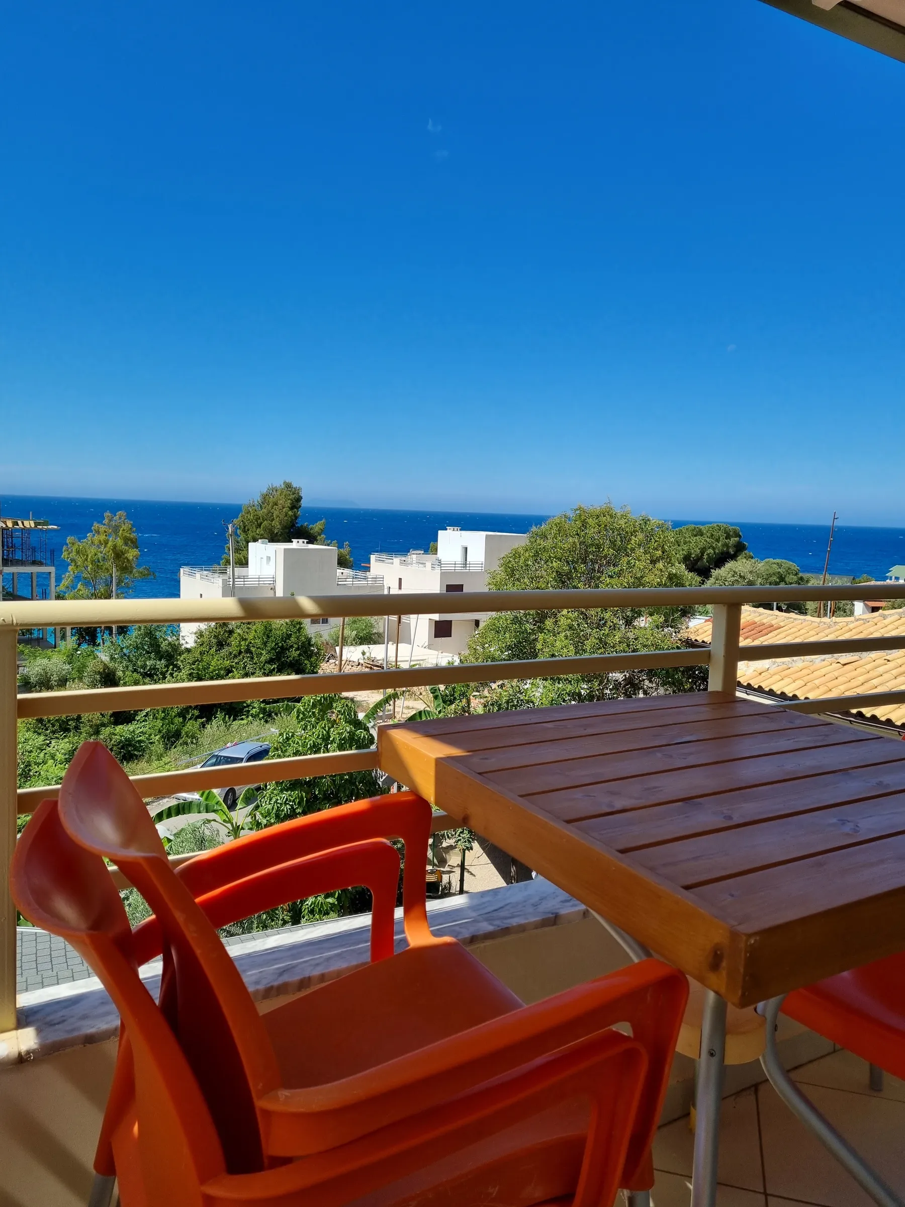 Orange chairs and a slatted timber table on a balcony at Azure Hotel Dhermi, with olive trees, white houses and the blue sea beyond the rail