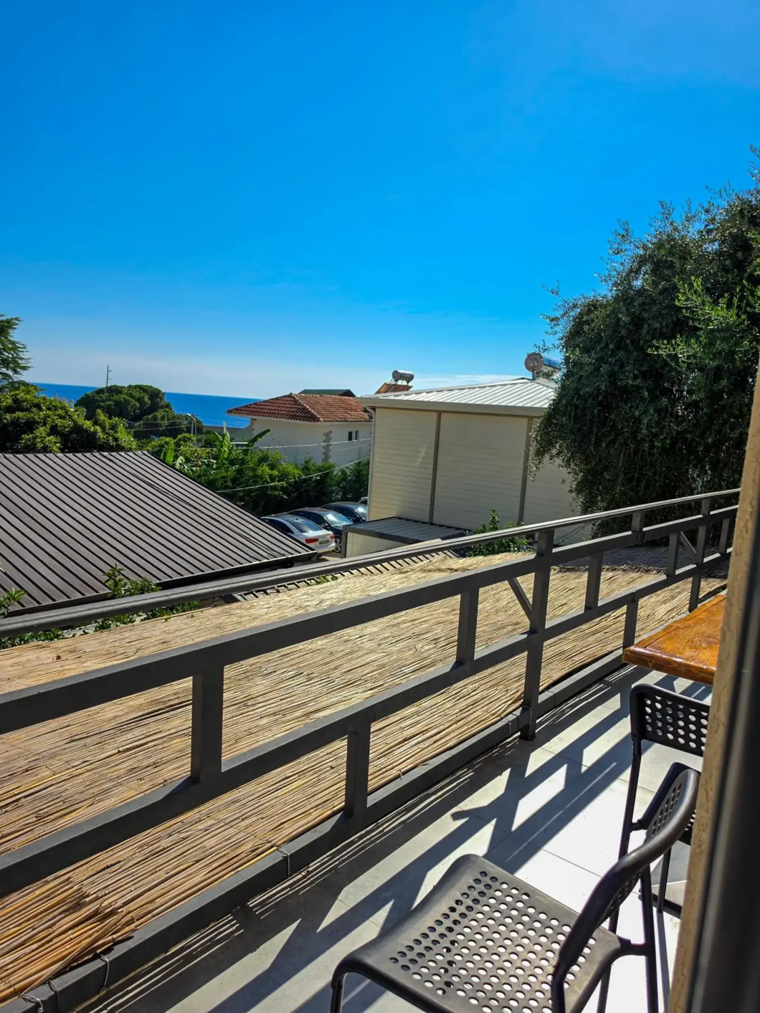An upper balcony with a dark rail and a reed screen, looking over grey metal roofs and trees to a line of blue sea