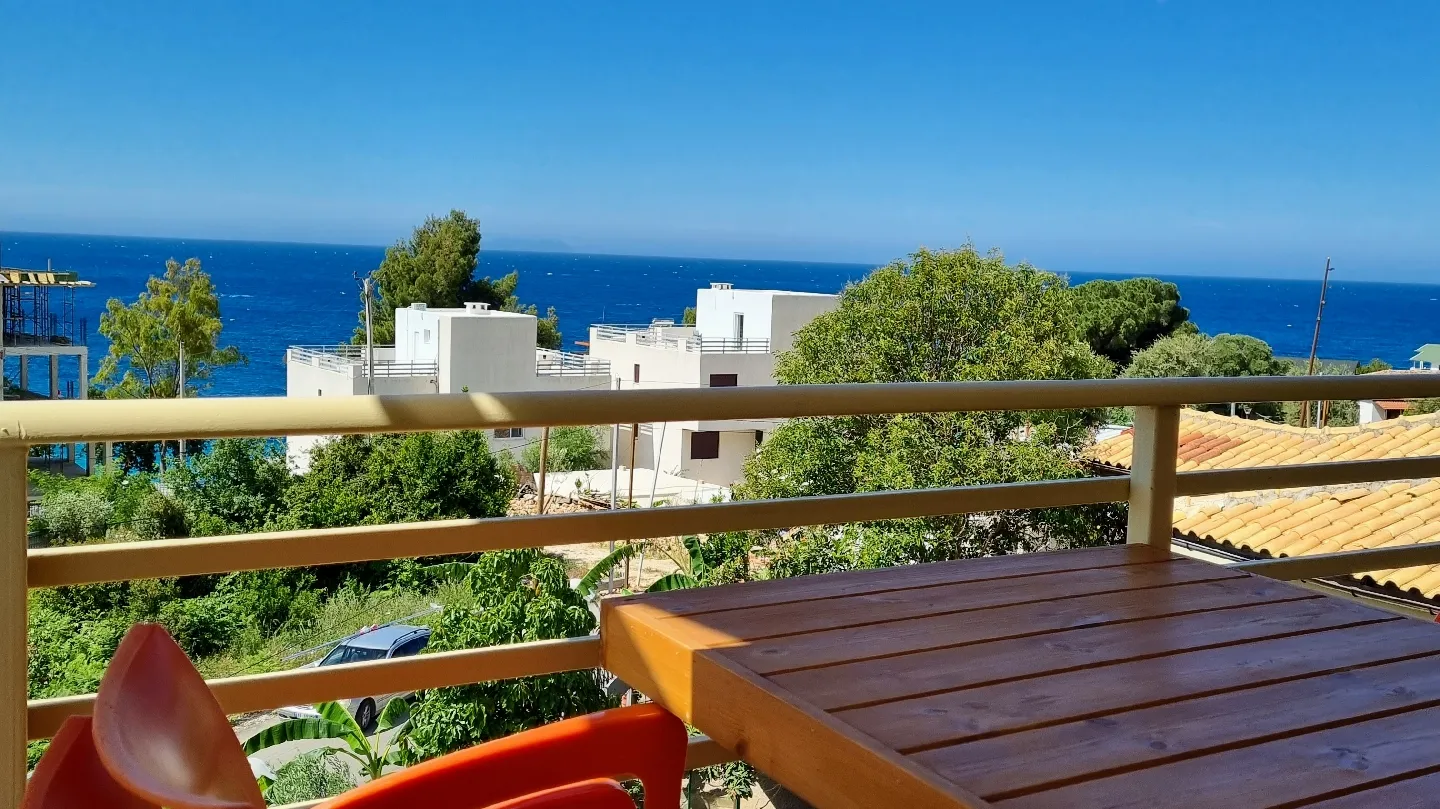 A timber balcony table above garden trees and tiled roofs, with white houses and the open sea under a clear sky