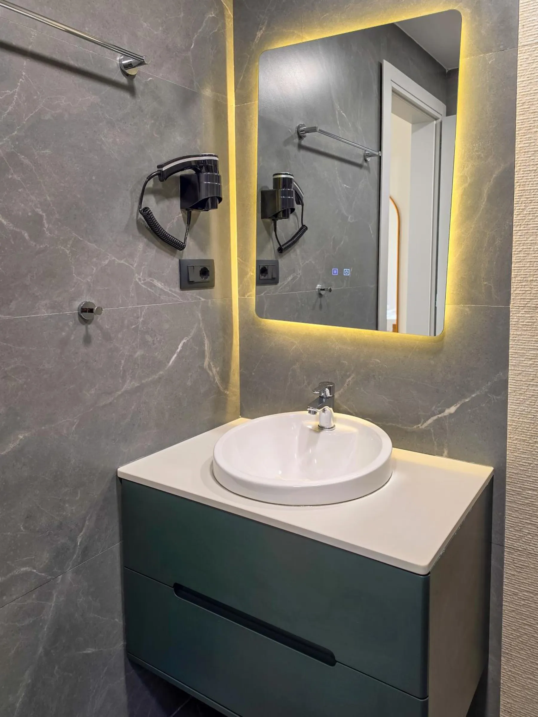 A round white basin on a dark green vanity under a mirror lit from behind, with a hairdryer on grey stone-effect tiles