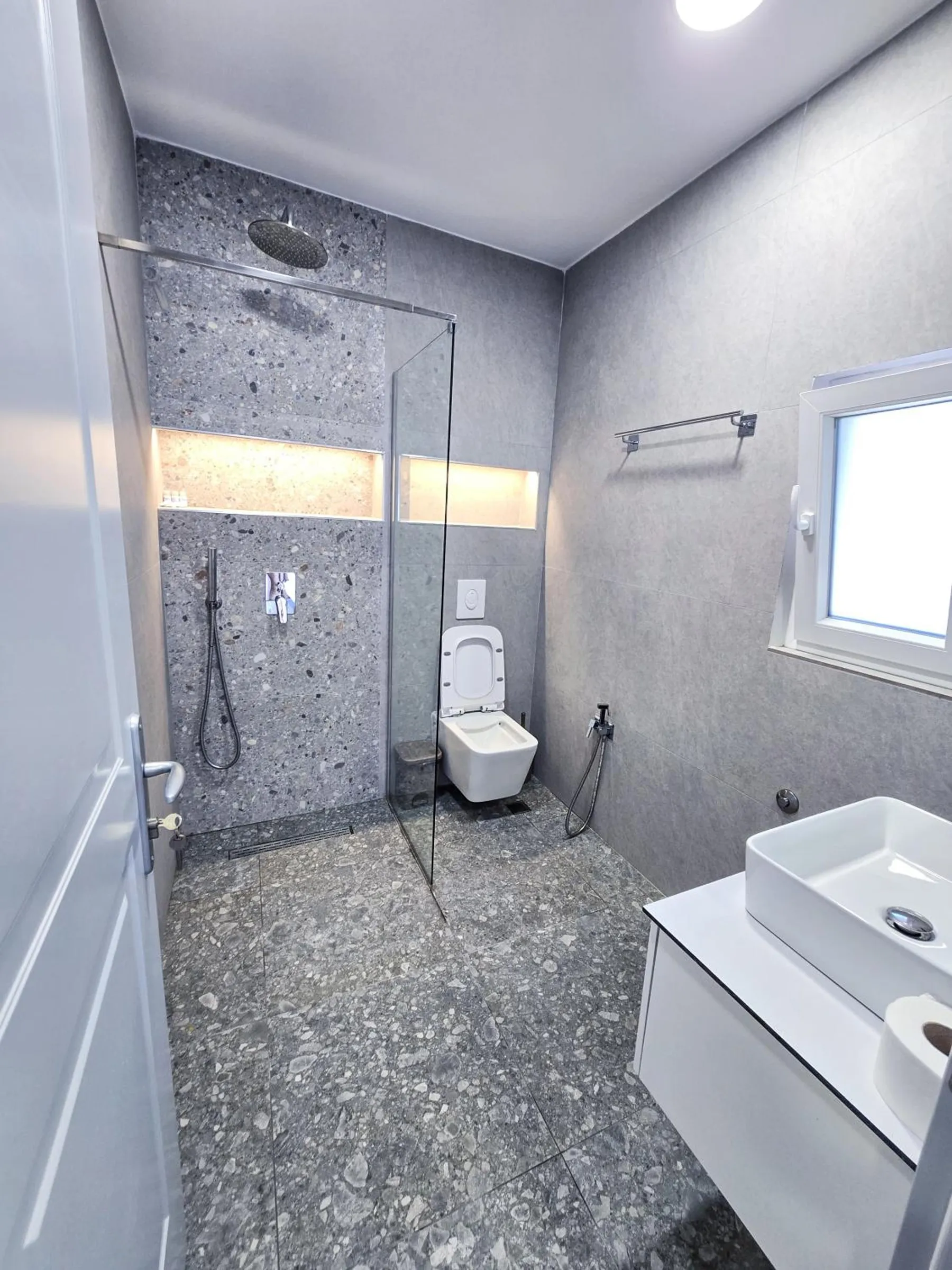 A shower room with a terrazzo floor and wall, a glass screen, a lit niche, a wall-hung toilet and a white vessel basin