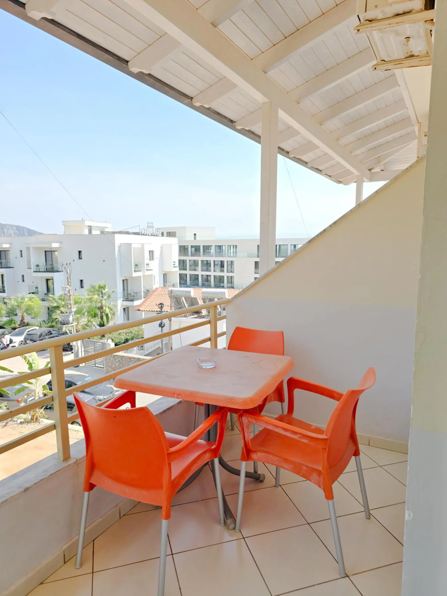 A covered balcony with a white timber ceiling, an orange table and three orange chairs, looking across to white buildings and the hills
