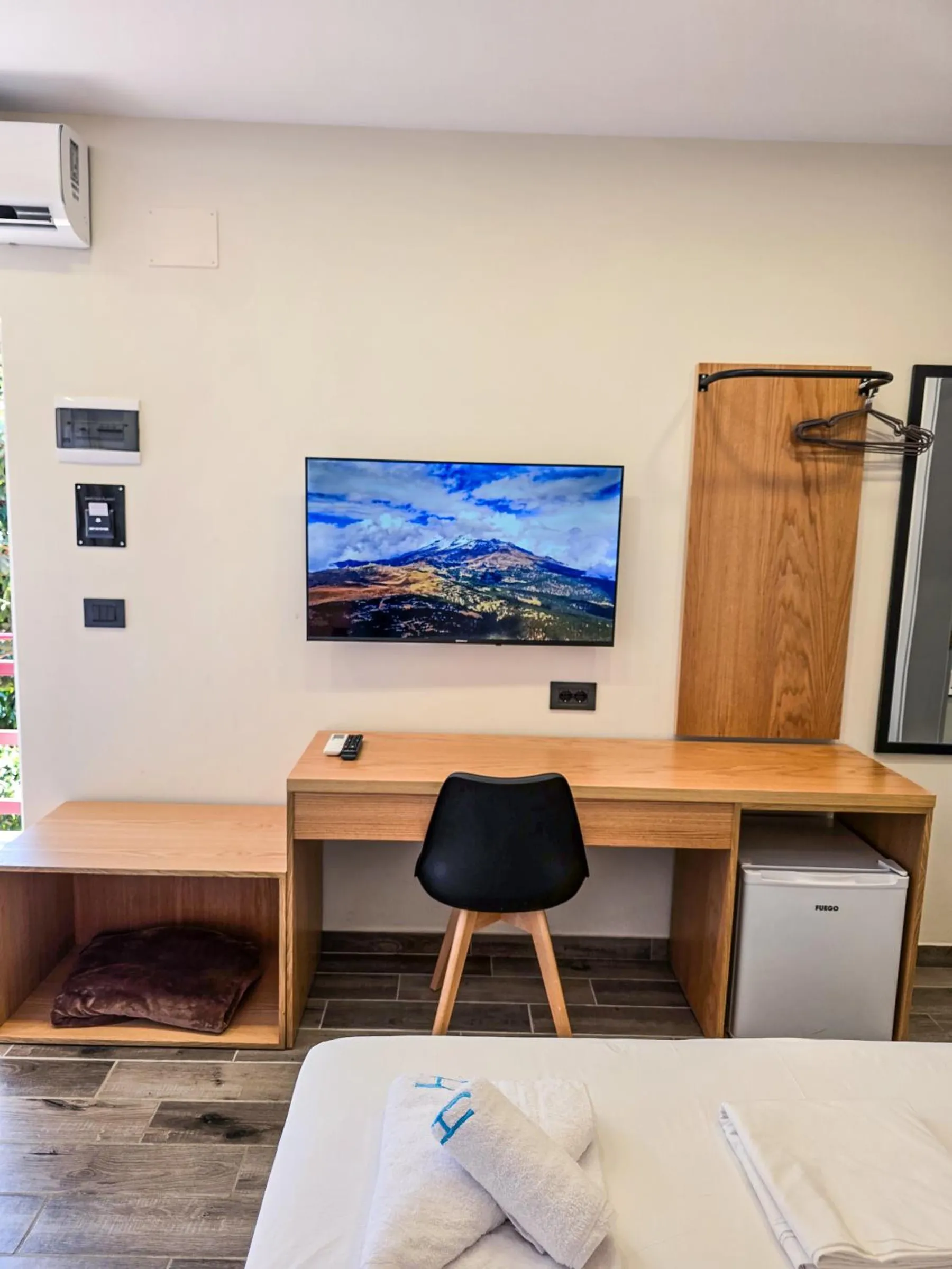An oak desk with a black chair, a small refrigerator underneath, a television above and an oak coat hanger by the door