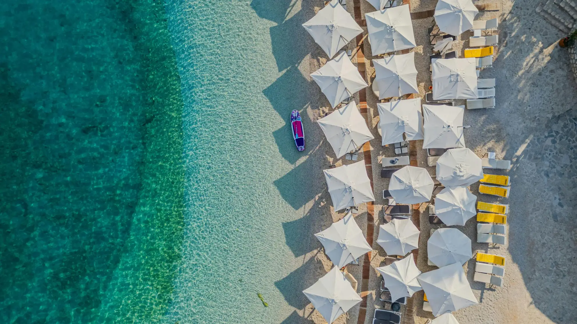 Dhërmi seen from directly above: turquoise water over pale pebbles and rows of white umbrellas and loungers on the beach