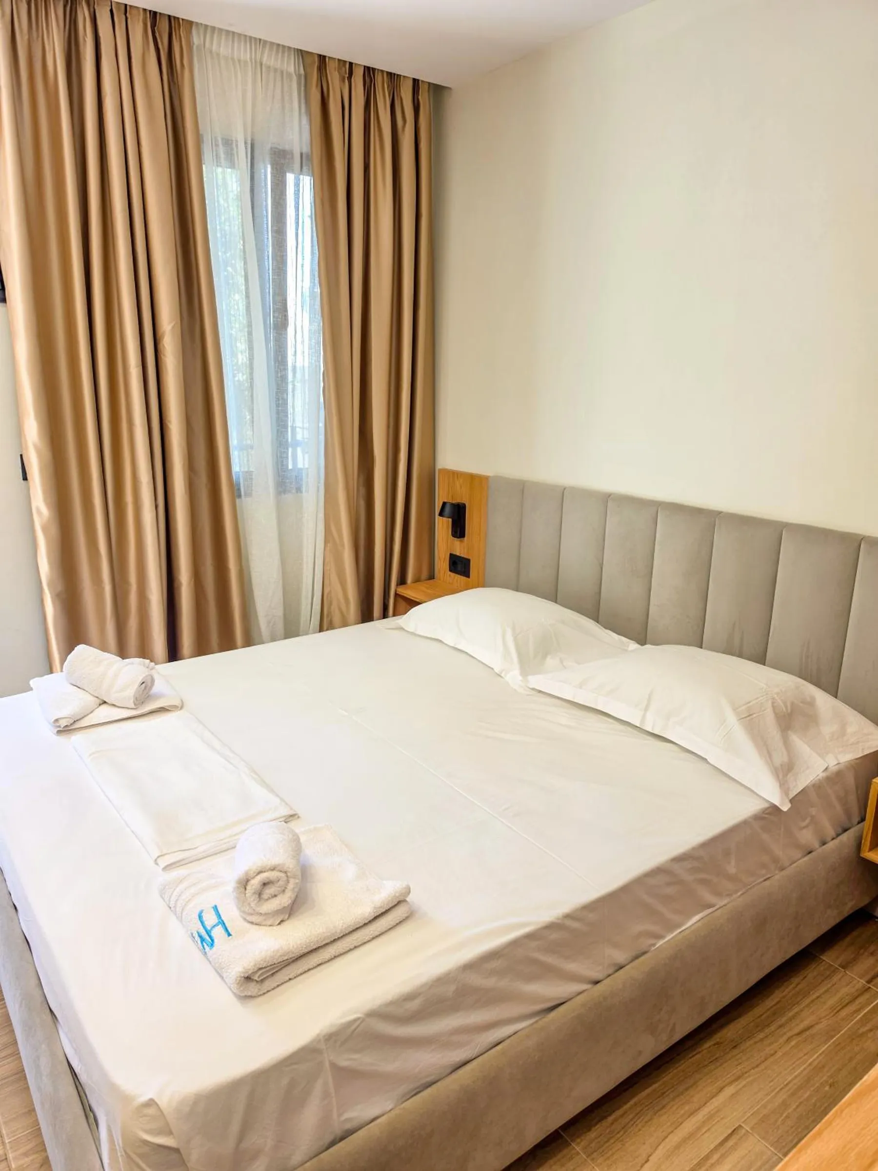 A double bed with white linen and a grey channelled headboard beside a window with gold curtains and sheers