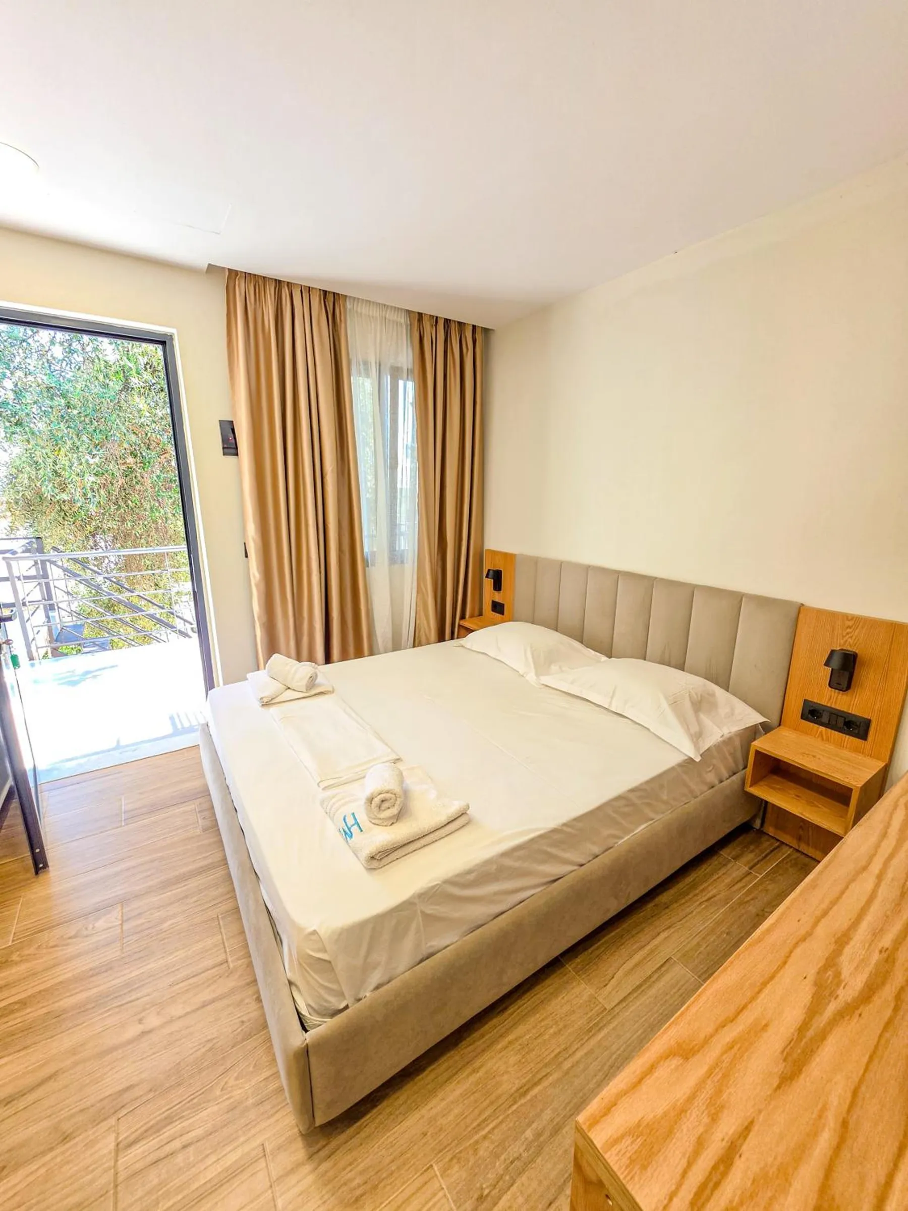 A double bed in an oak-and-grey room with a glazed door open to a balcony and trees outside
