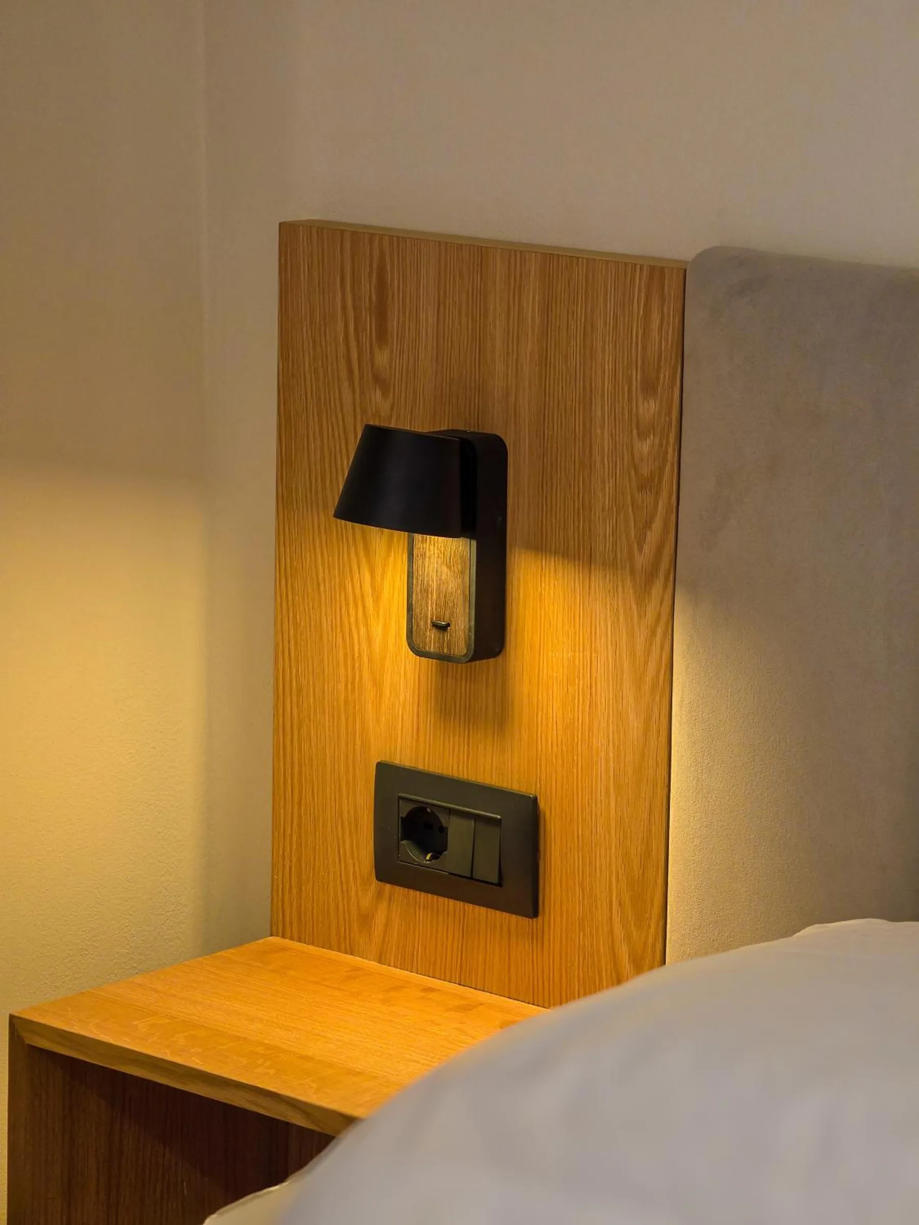 A black reading lamp and a pair of sockets on an oak panel beside a bed, with an oak shelf below