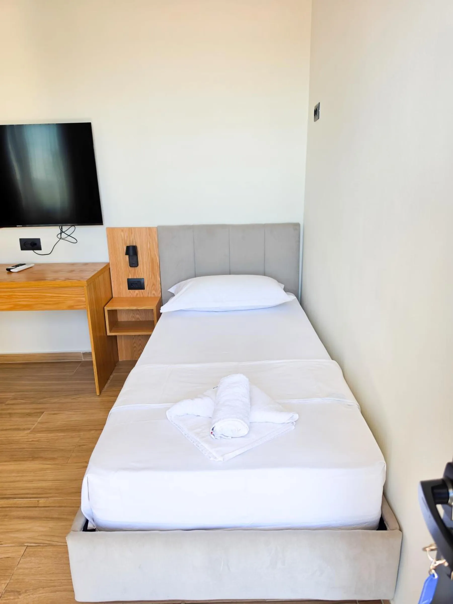 A single bed with a grey headboard and white linen, an oak desk and a television on the wall