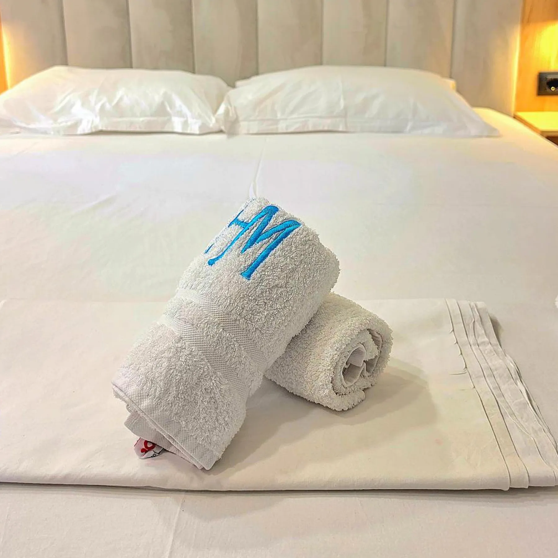 Two rolled white towels on a made bed, the front one embroidered with a monogram in azure thread