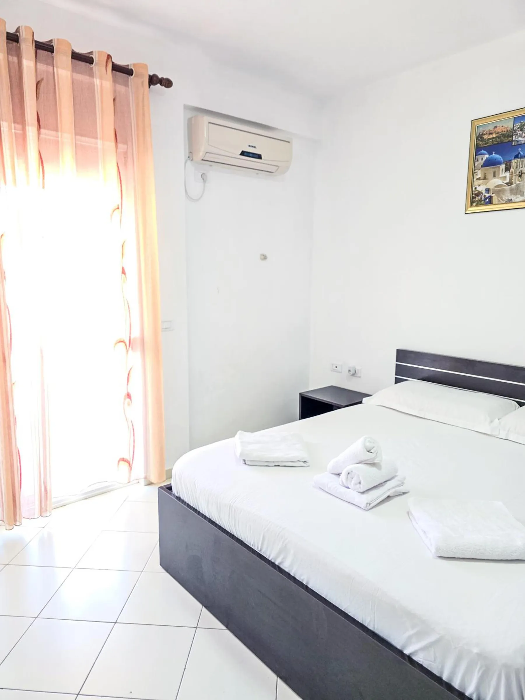 A double bed with a dark timber frame on a white tiled floor, pink sheer curtains at the window and an air conditioner on the wall