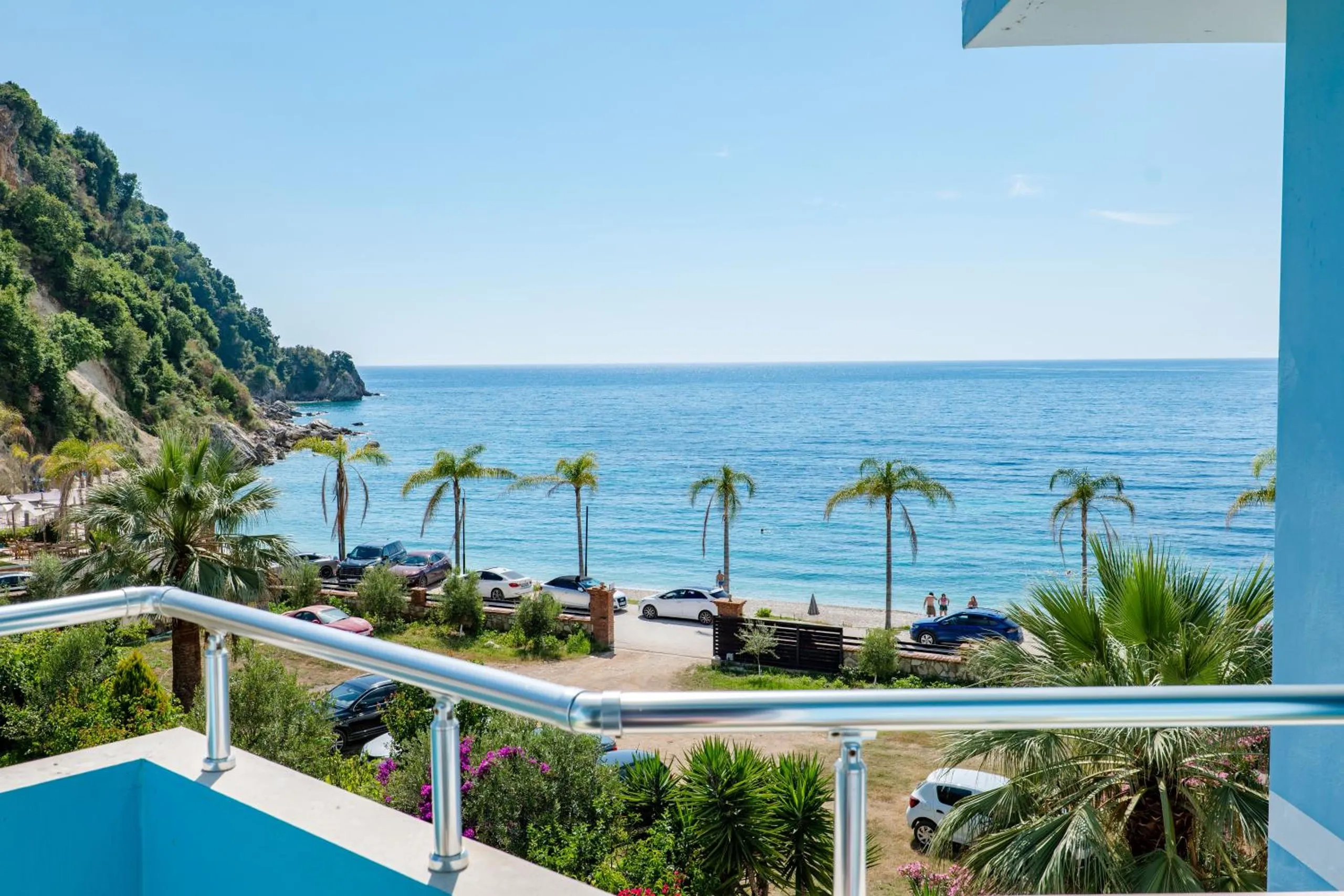 The view over a chrome balcony rail: palm tops along the promenade, the green headland on the left and a flat calm sea