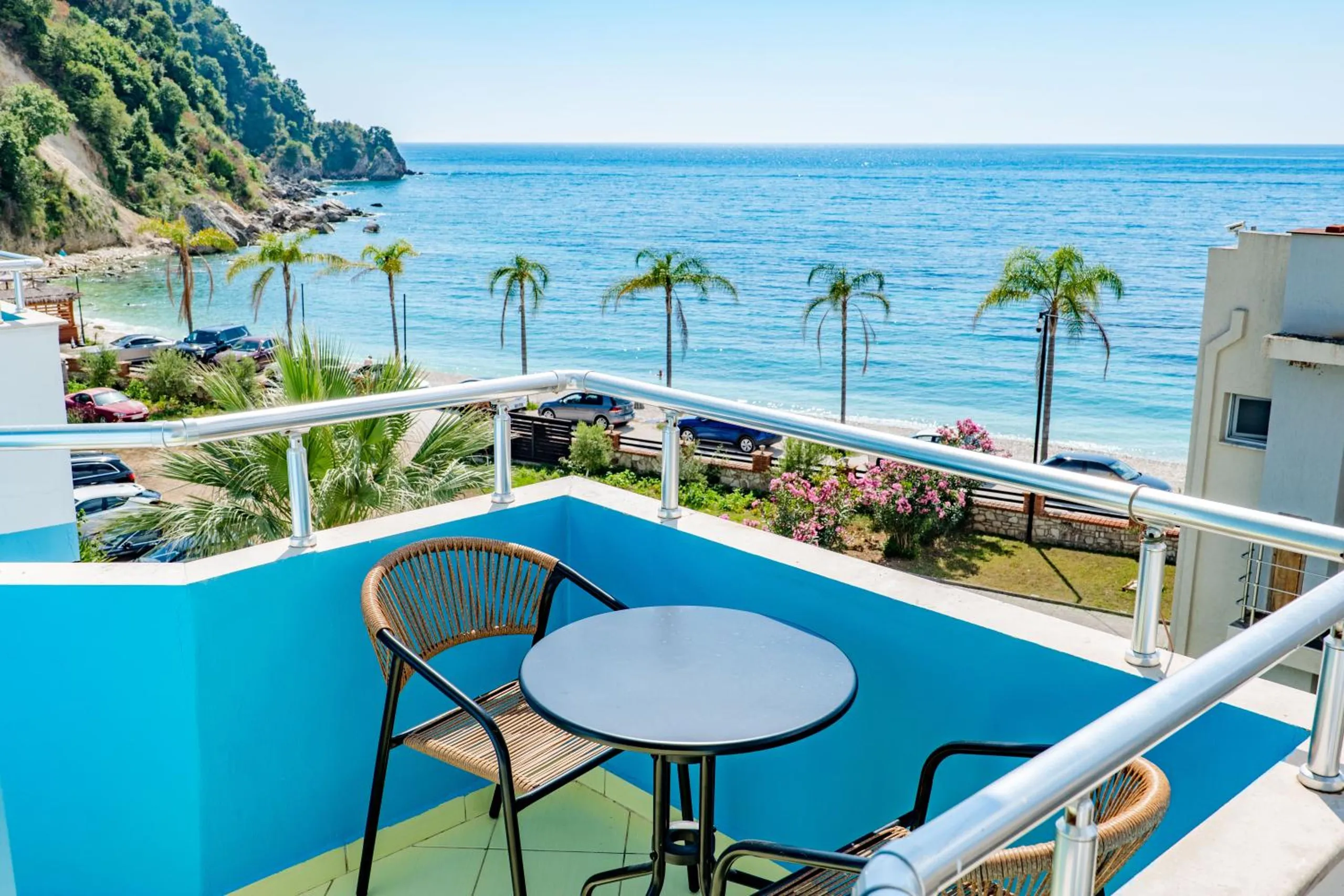 A round table and two rattan chairs on a balcony painted cerulean, a chrome and glass rail, and below it palms along the promenade, Potam Beach and the open sea under a clear sky