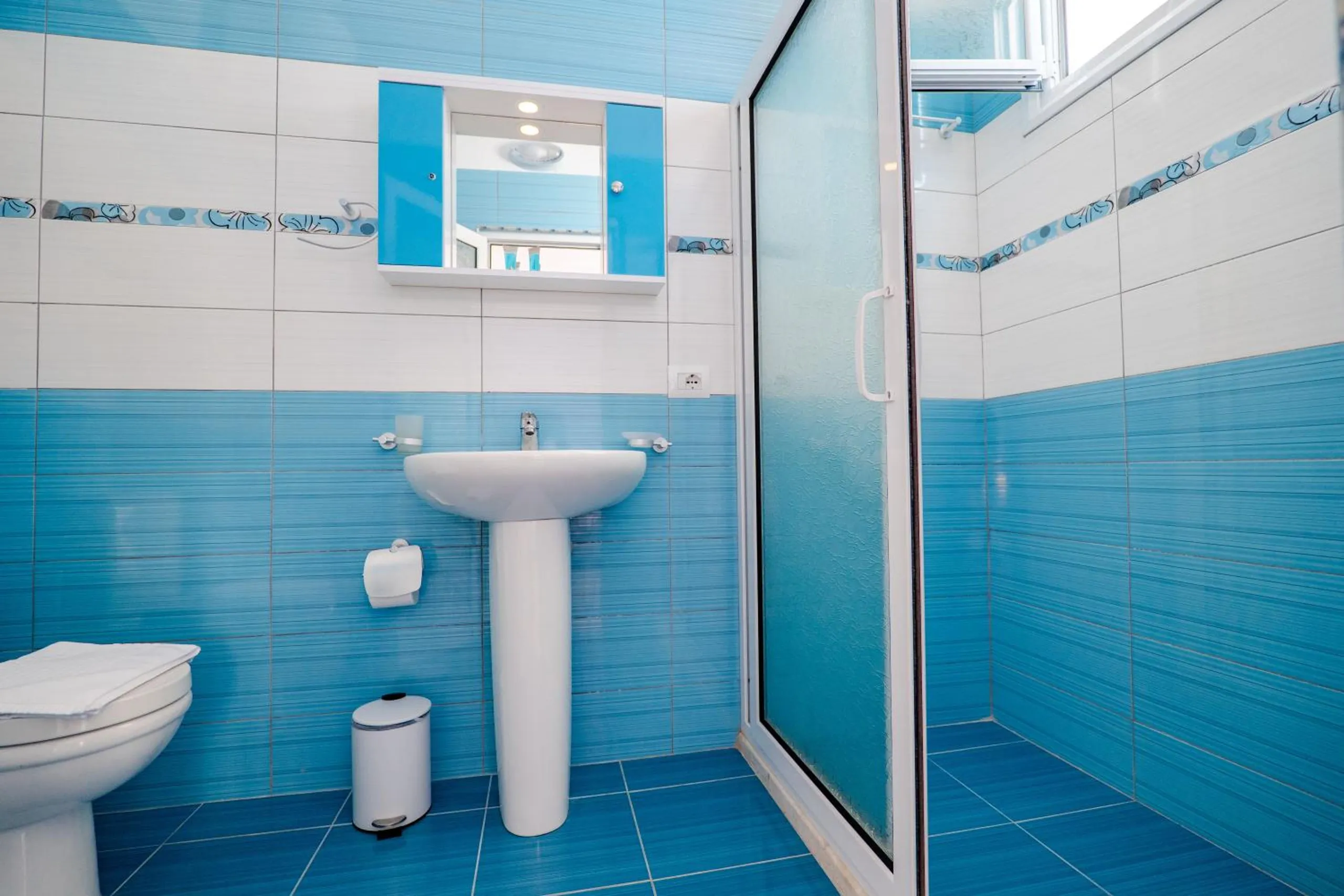 A bathroom tiled in bright blue and white with a pedestal basin, a toilet and a glass shower cubicle