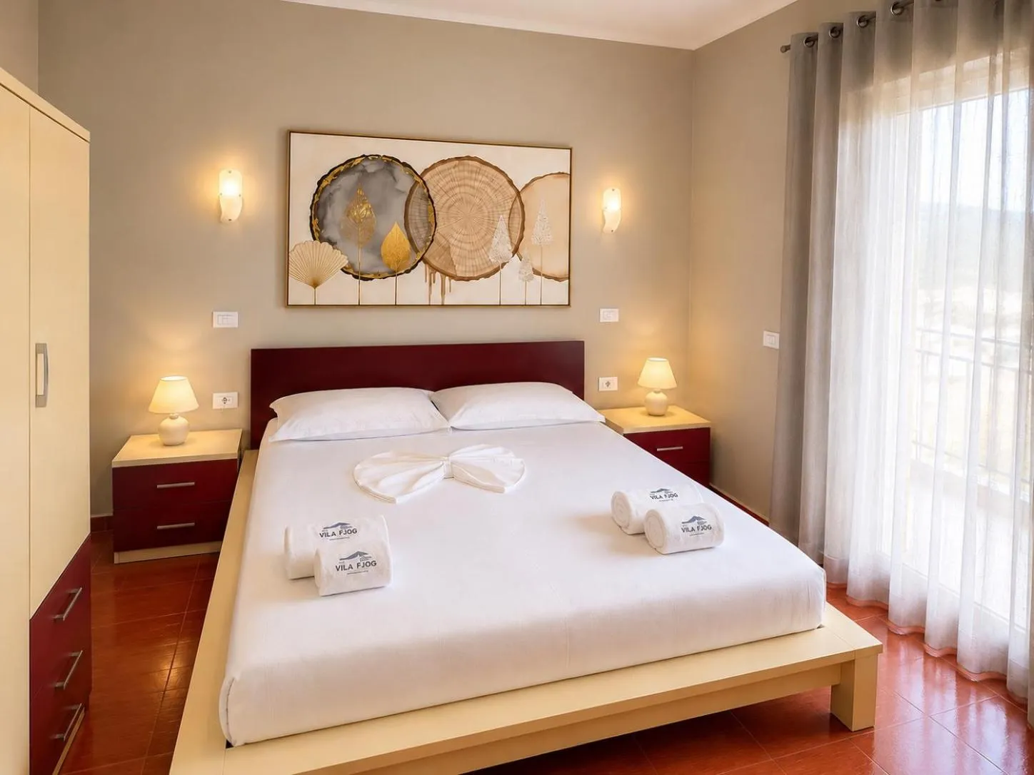 A double bed with a burgundy headboard, warm wall lights either side and long sheer curtains over the balcony door