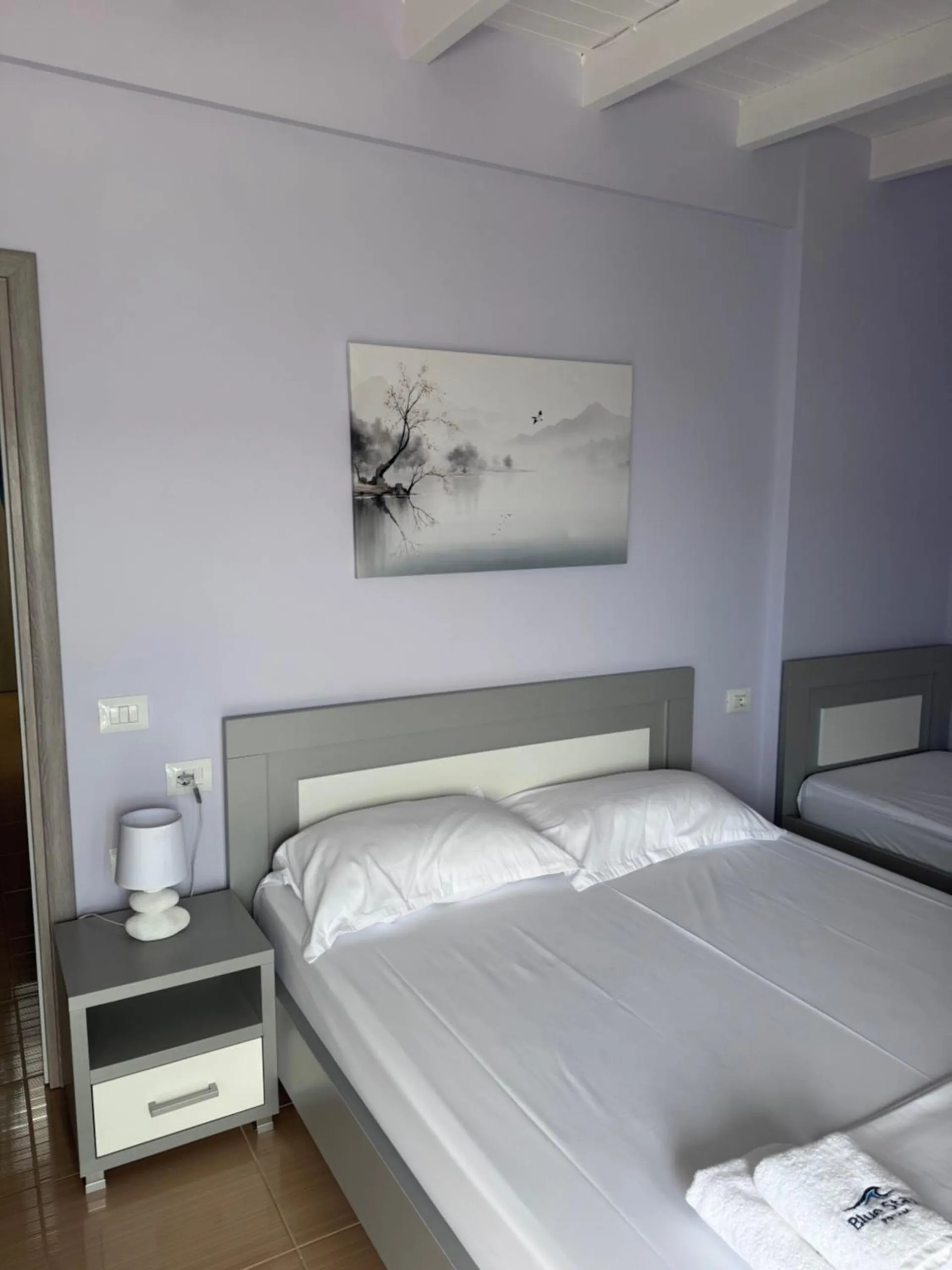 A double bed with a grey-white frame against lilac walls, a white bedside table and lamp, an ink-wash print above