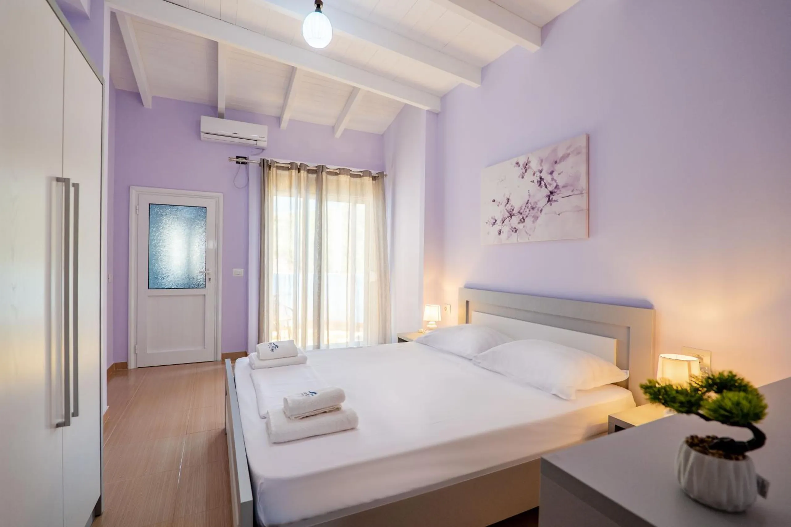 A double room with lilac walls and a white beamed ceiling, a grey-framed double bed with folded towels and a bonsai on the bedside table