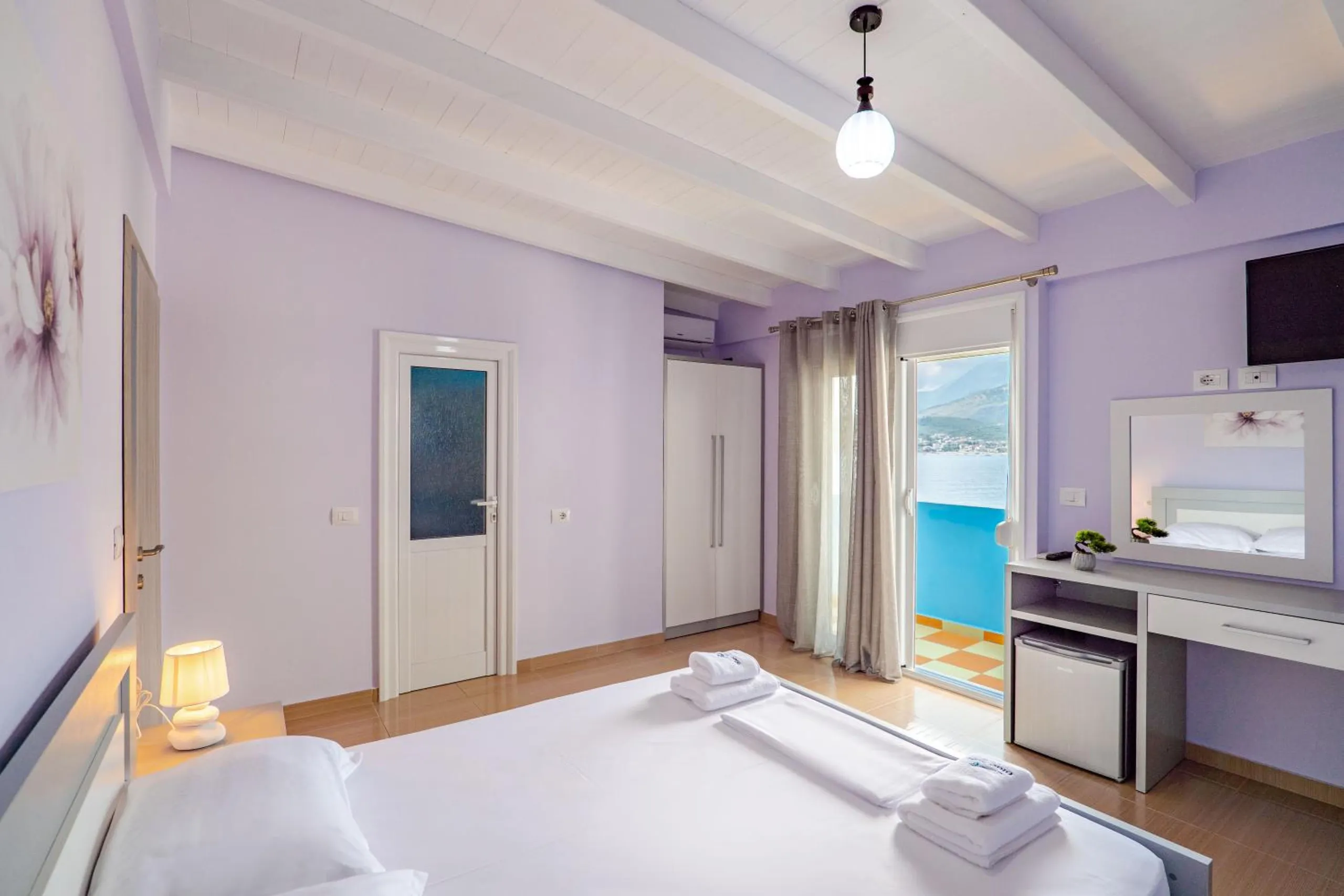 A lilac room with a double bed, a dressing table and an open balcony door showing the cerulean balcony and the sea