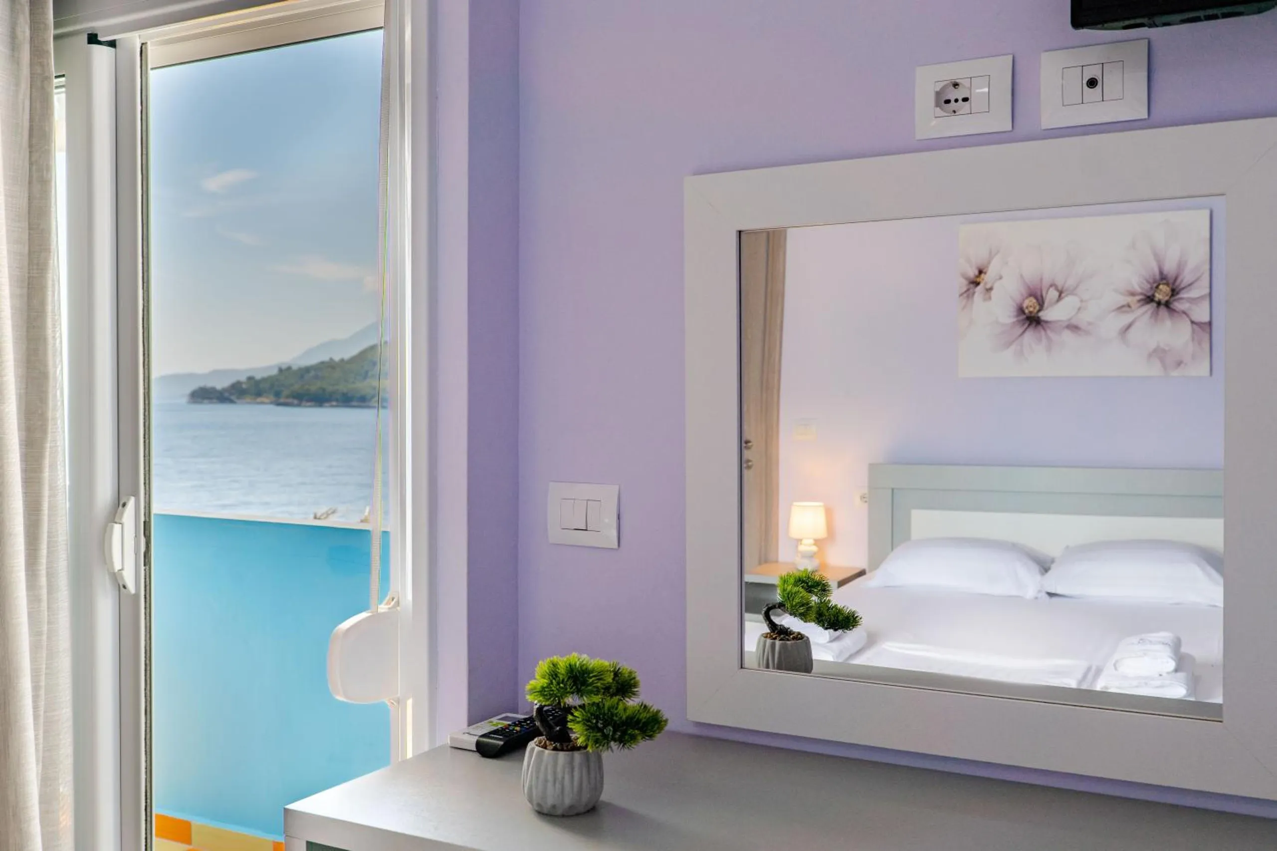 A mirror over a white dresser reflecting a made double bed and blossom prints, the balcony and the sea through the glass door to the left