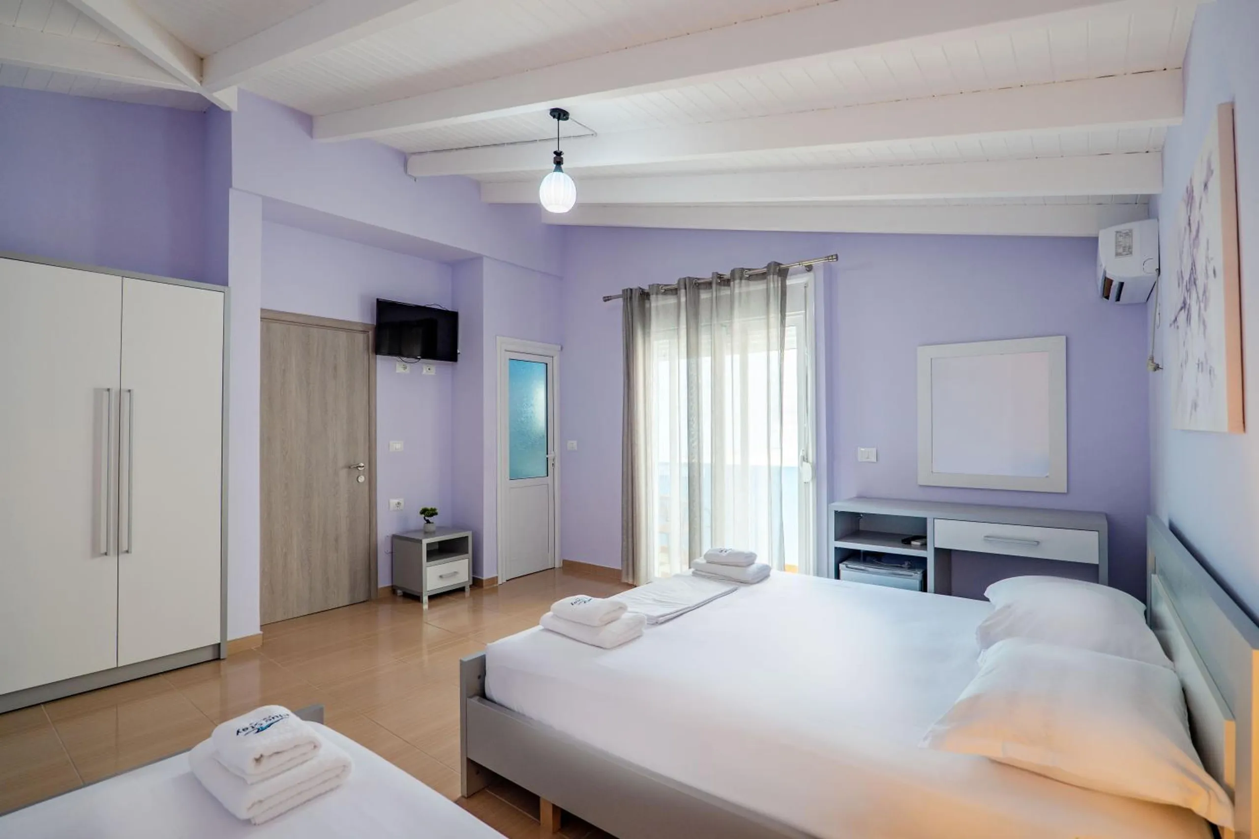 A lilac room with a double bed, a single bed, a tall white wardrobe, a desk with a mirror and a television on the wall