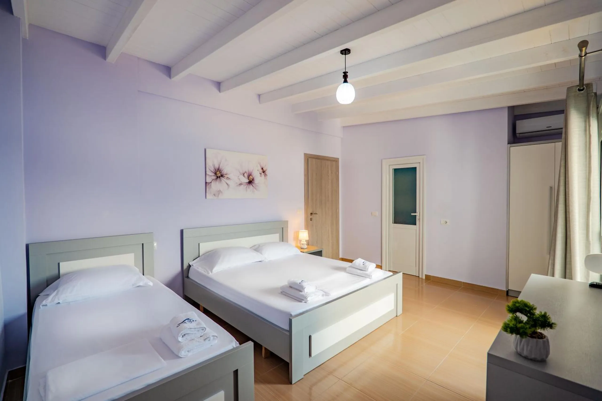 A lilac room under white beams with a double bed and a single bed side by side, a balcony door hung with sheer curtains