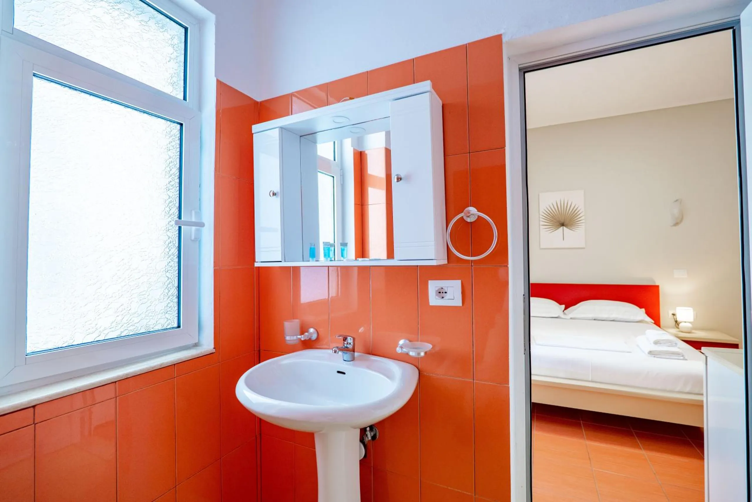 An orange-tiled bathroom with a white basin and mirror cabinet, the bedroom and a red bed visible through the open door