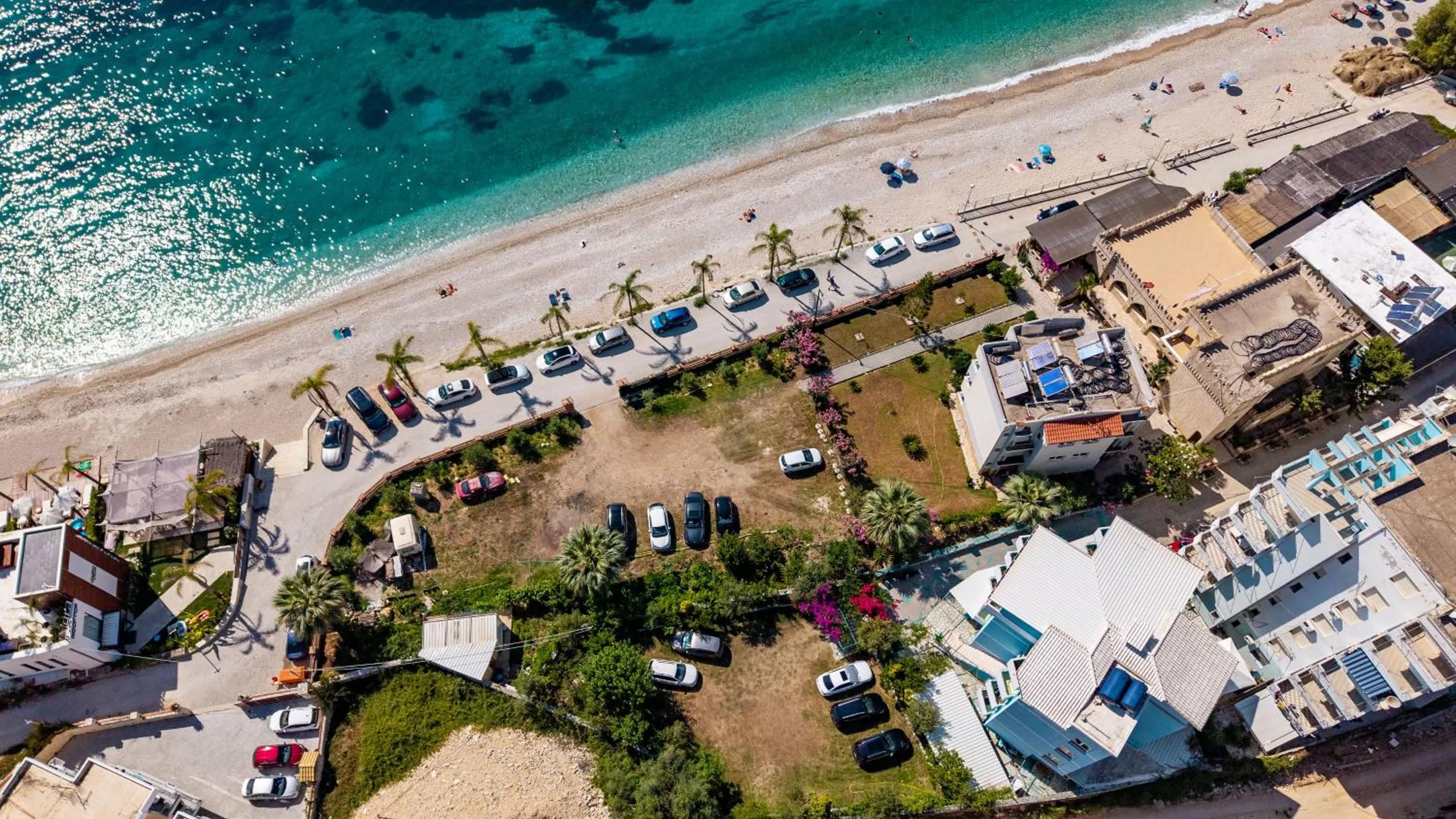 An aerial view along Potam Beach: clear shallow water, the shingle, a row of palms and parked cars on the promenade, and the garden and white roofs of the houses behind