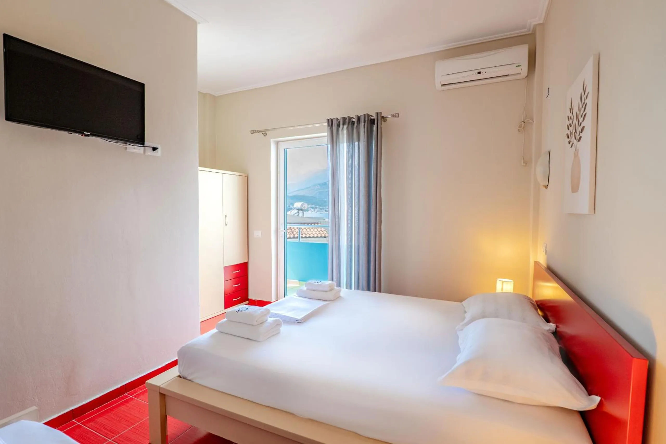 A double room with a red headboard and red floor tiles, the balcony door open to blue railings and the sea
