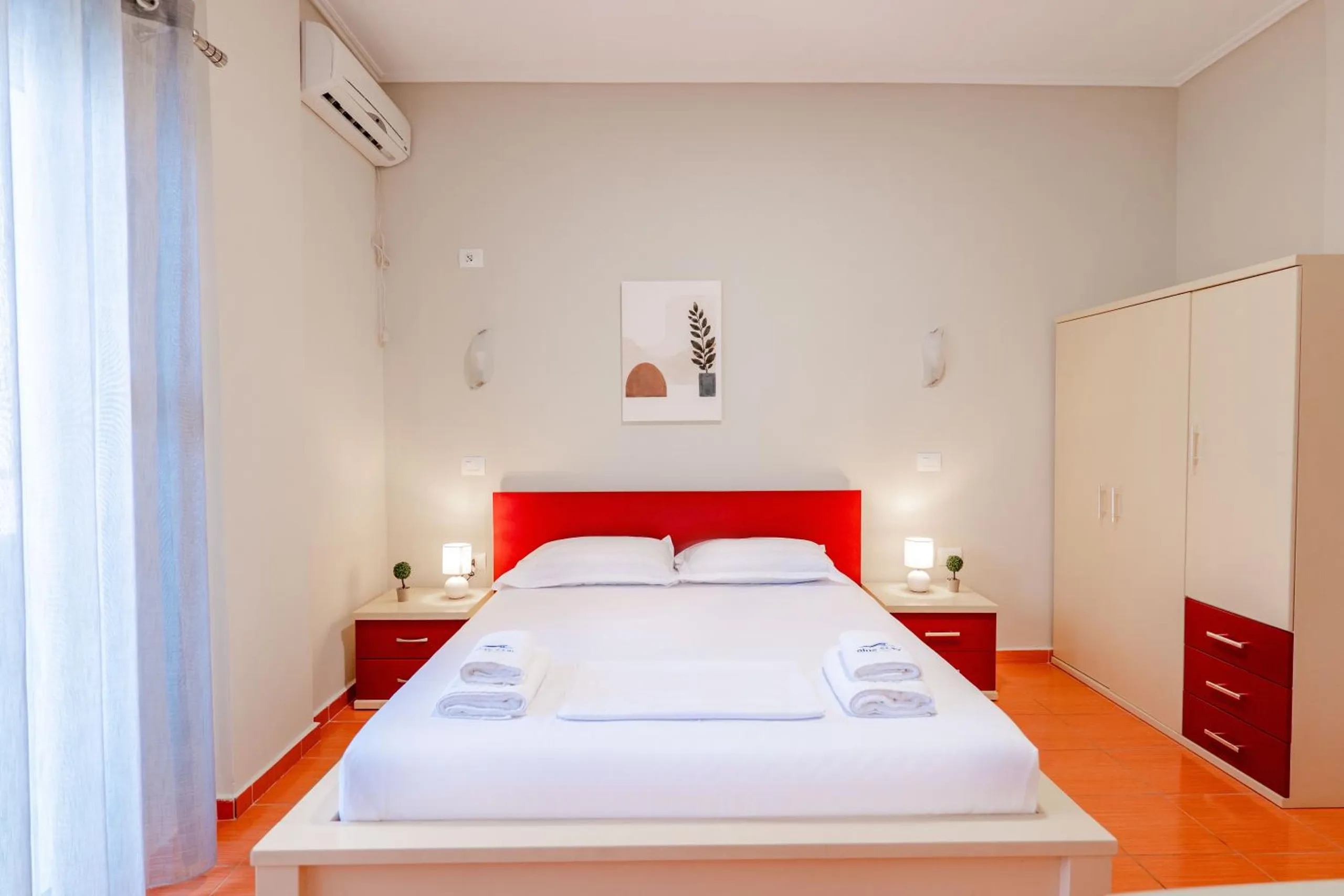A double bed with a red headboard on an orange tiled floor, a cream wardrobe with red drawers and a leaf print above the bed