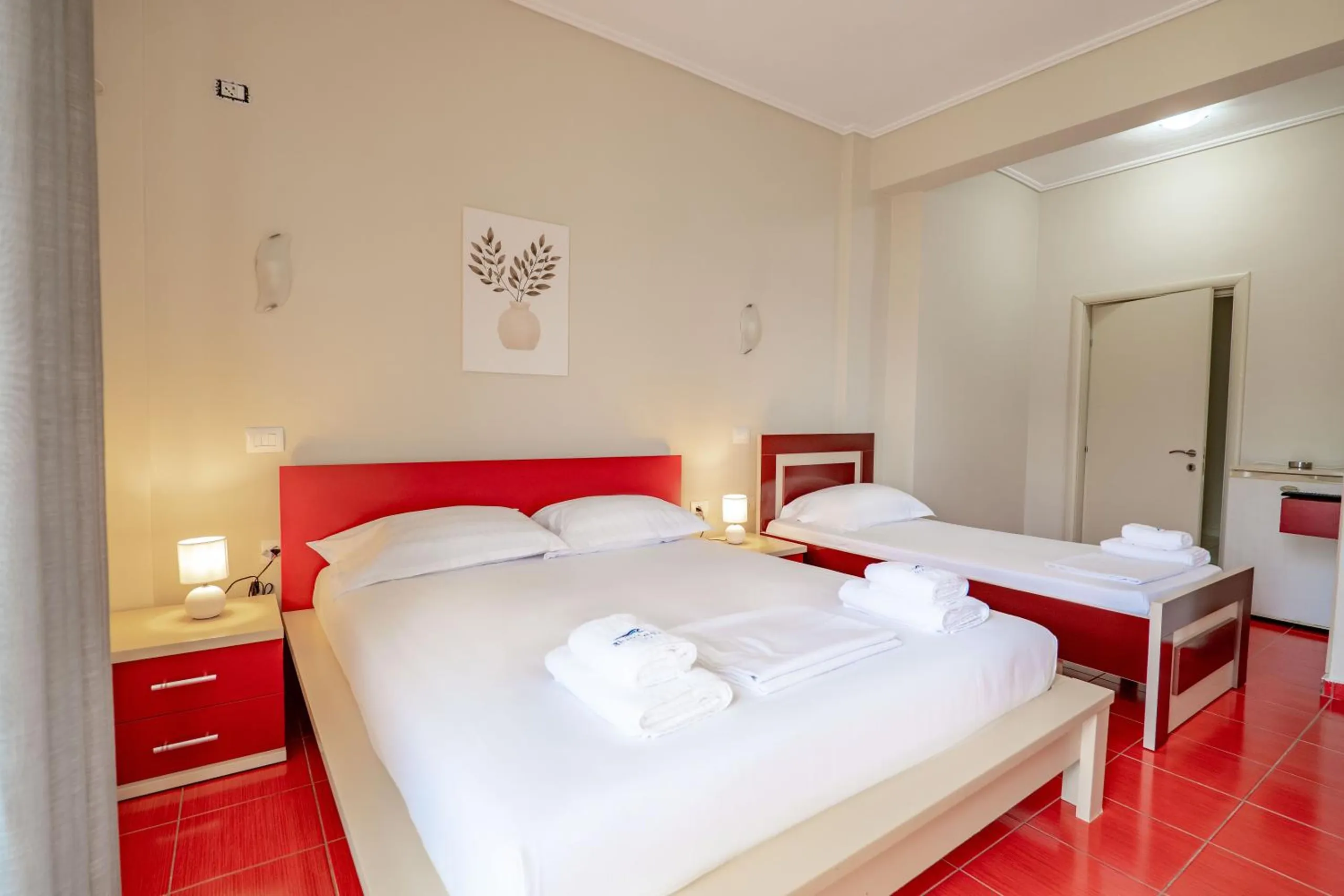 A room with a double bed and a single bed, both with red lacquered frames, red bedside tables and two lamps lit against cream walls