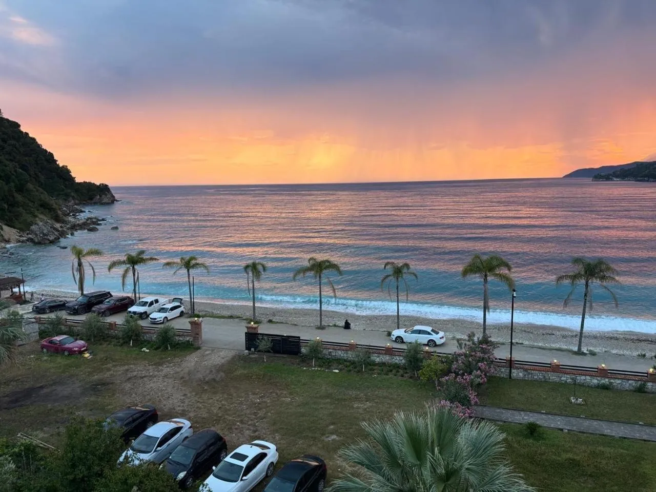 A pink and orange sky over Potam Beach, the palms along the promenade and a few cars parked under them