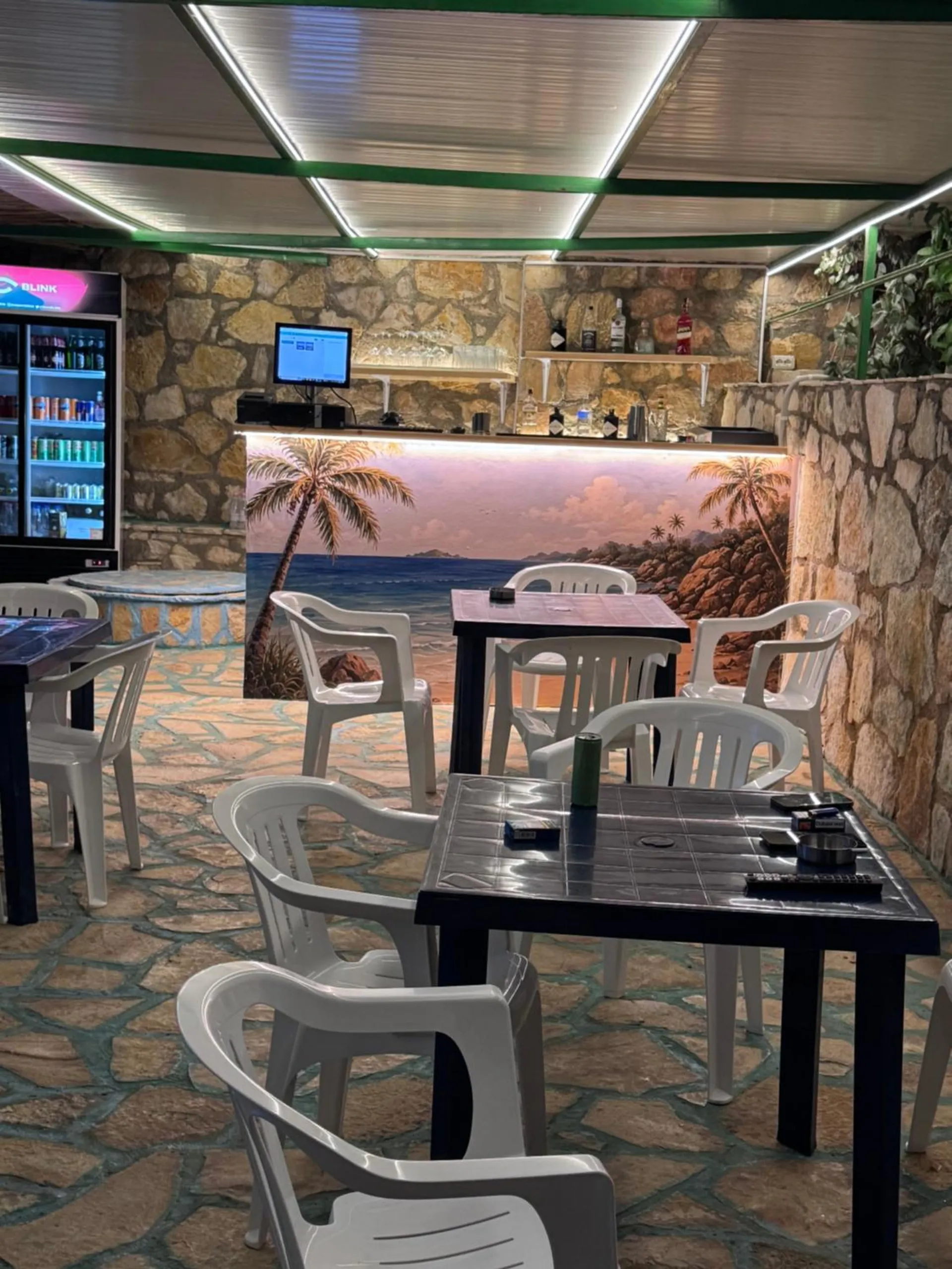 The bar under a translucent roof: stone walls, a counter painted with palms and a beach, drinks fridges, and black tables with white chairs
