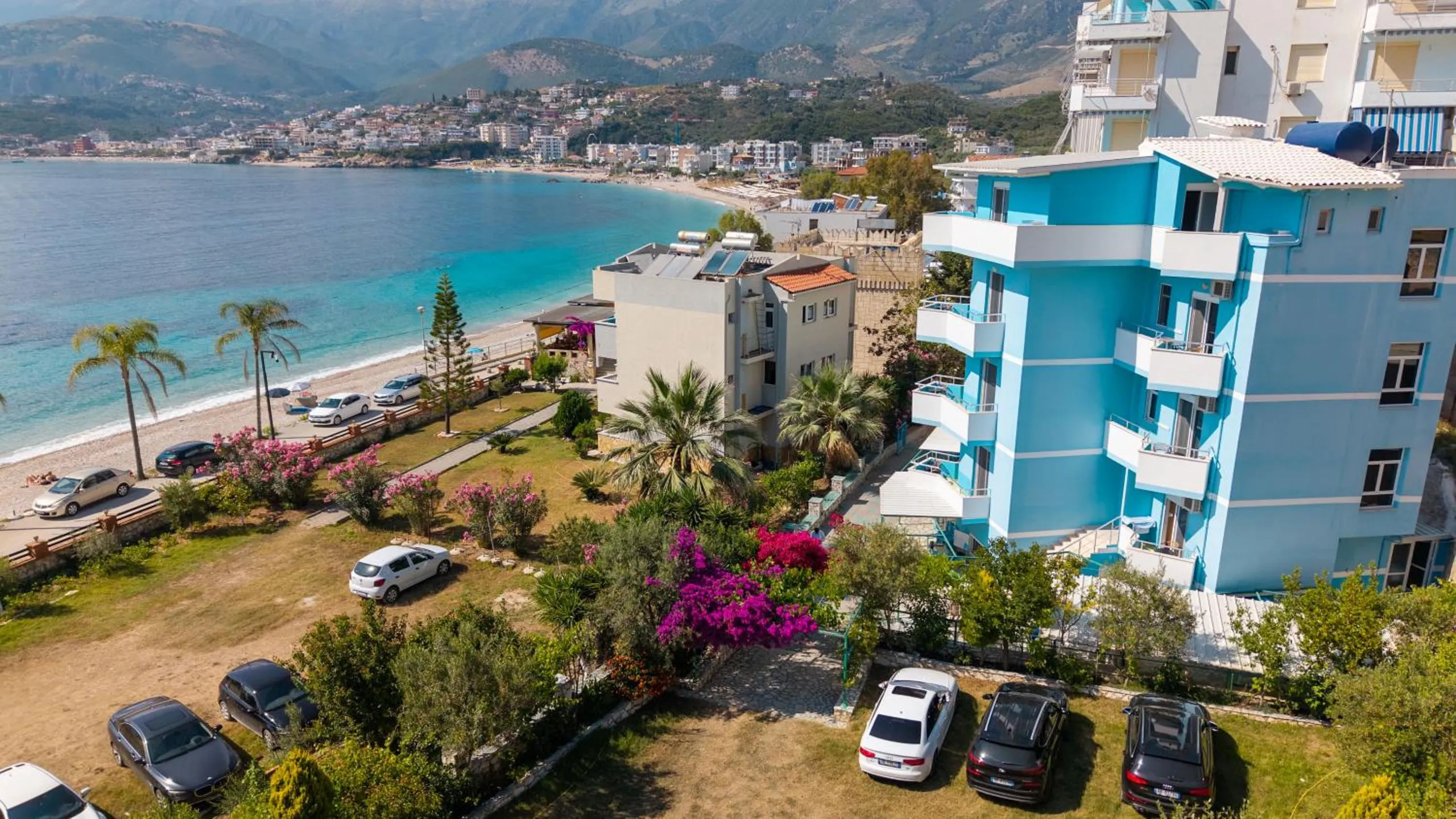 The hotel from the air: a five-storey building painted sky blue with white balconies, a garden of palms and pink bougainvillea in front, Potam Beach curving away towards Himarë town and the mountains
