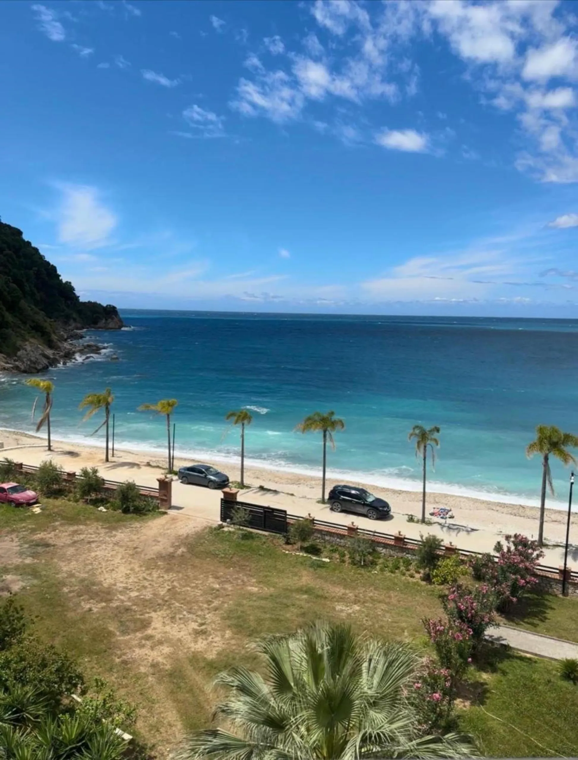 Looking down from an upper floor at midday: the garden, a row of palms, a white beach and turquoise water under scattered cloud