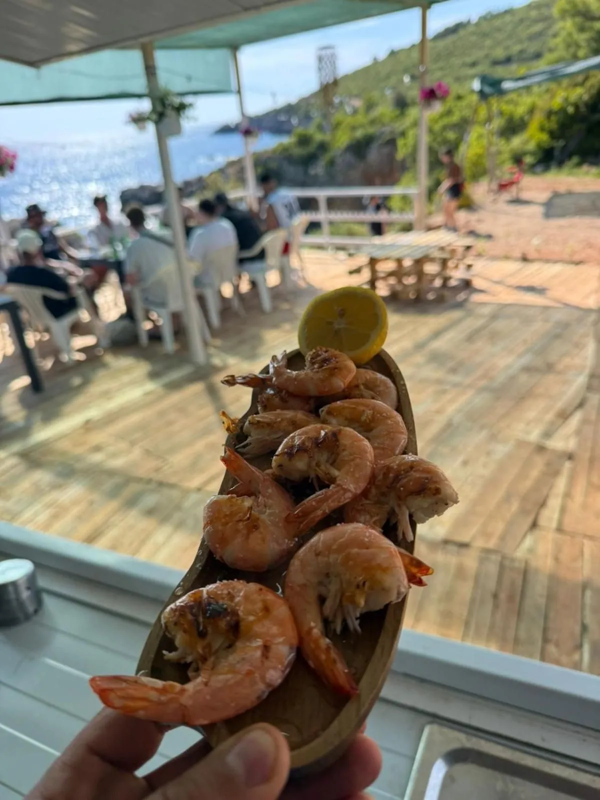 A wooden dish of grilled shrimp held up over the terrace, the sea and the headland behind