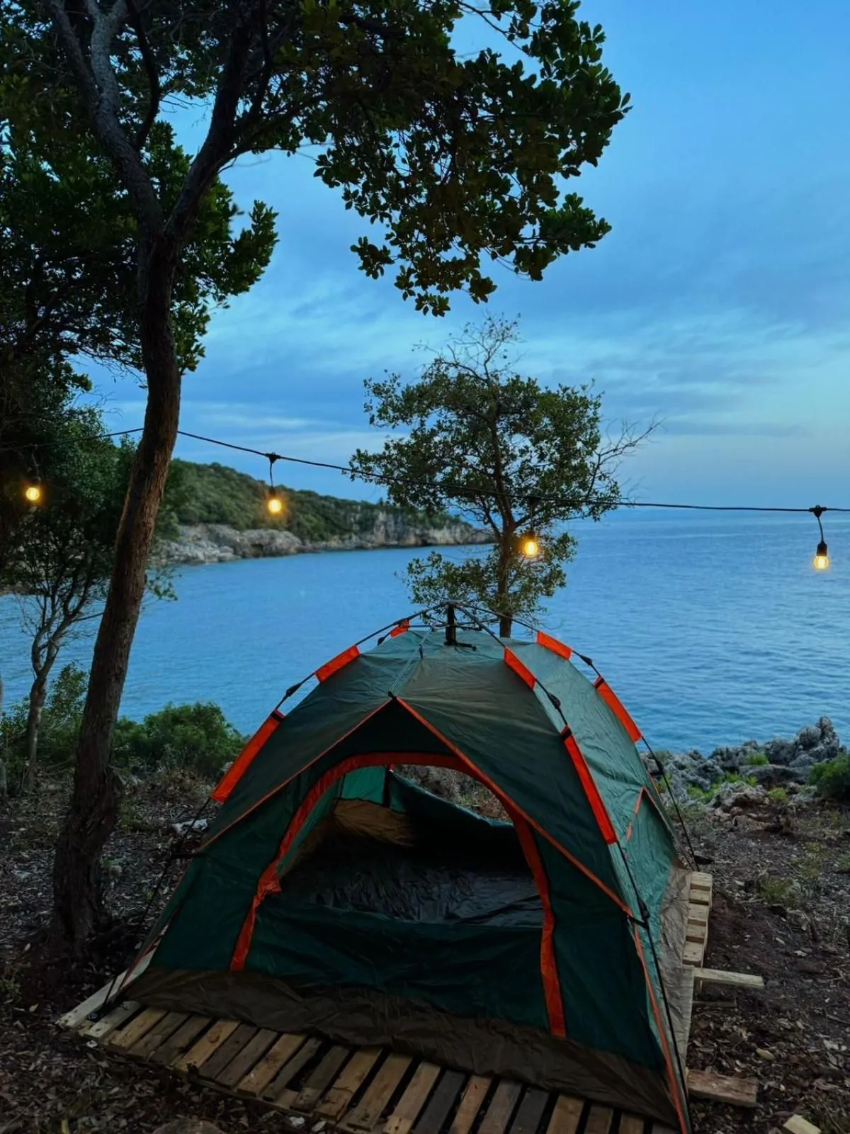 A dome tent on a pallet deck at the cliff edge, string lights overhead and the bay at blue hour