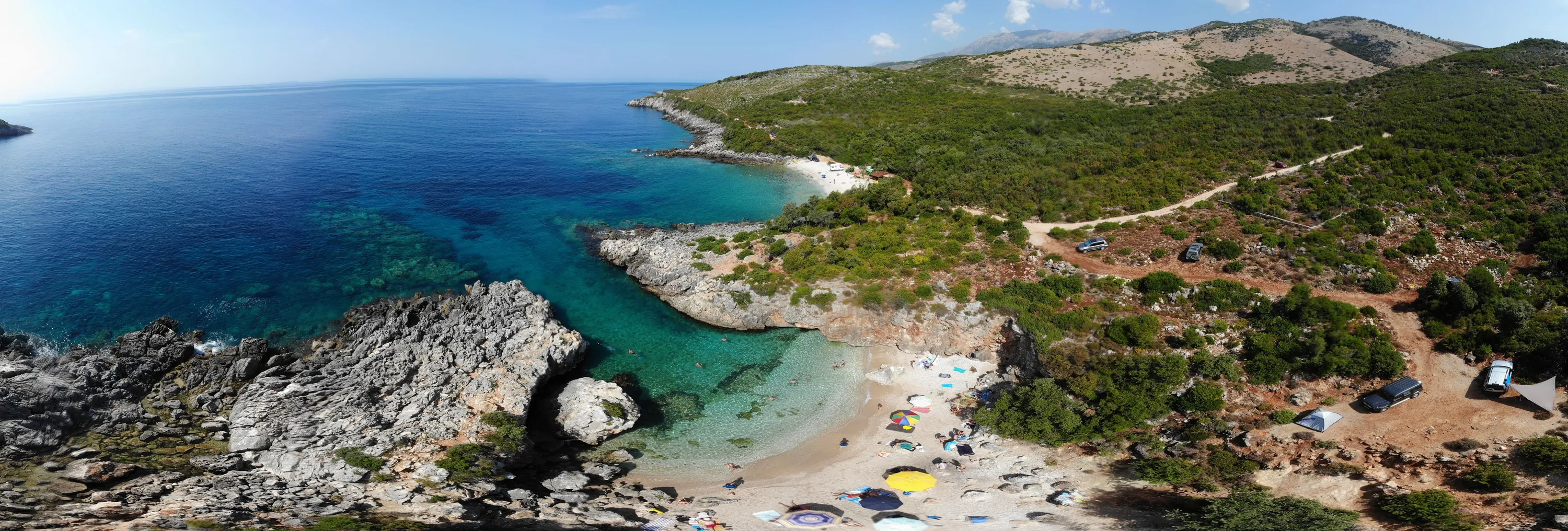 Aquarium Bay from the air: a small pebble cove of turquoise water between limestone rocks, a dirt track and wooded slopes above it
