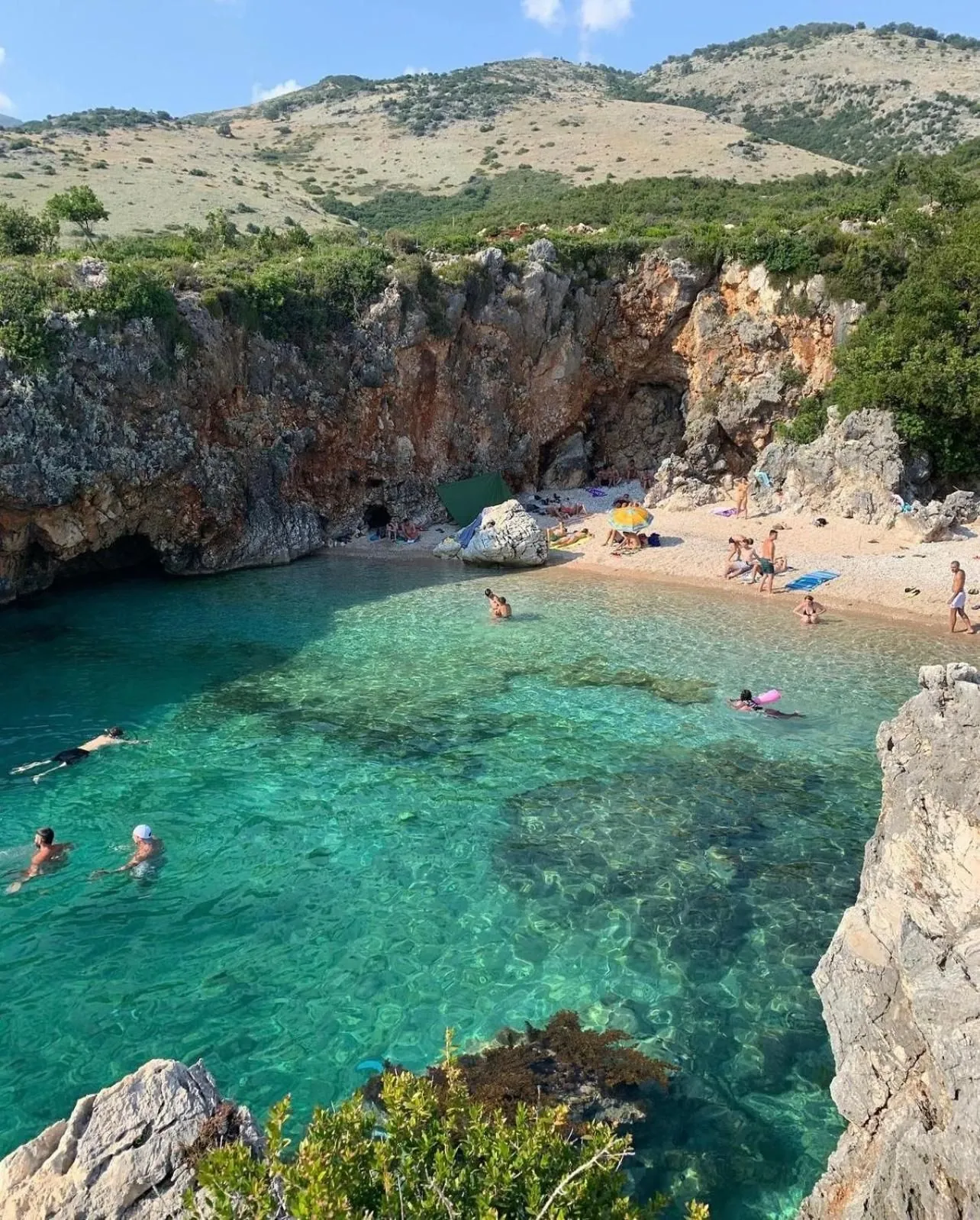 Swimmers in the clear green water of the cove, with a cave in the cliff behind the pebble beach