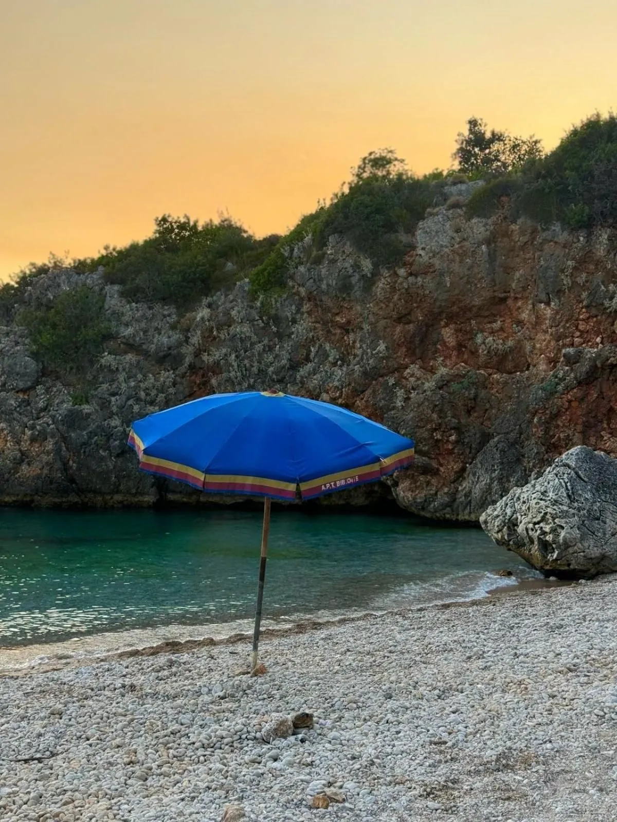 A blue umbrella planted on the empty pebble beach at dusk, red cliffs behind