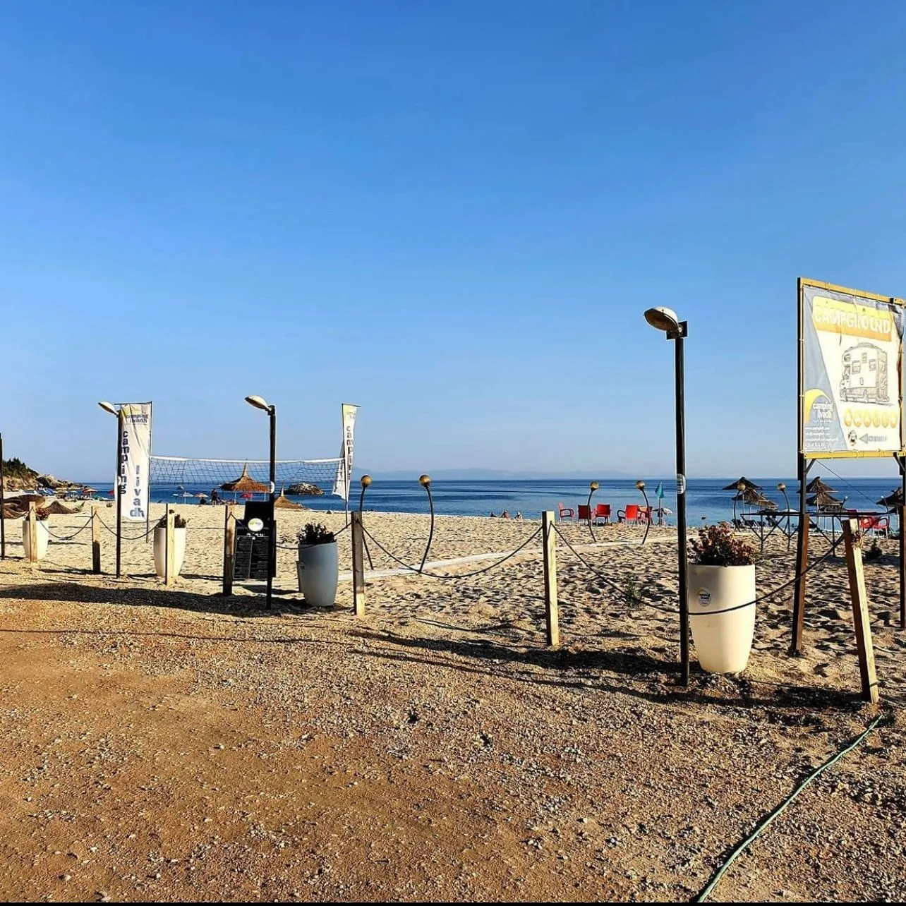The camp's way onto the beach: Camping Livadh banners, a volleyball net, rope-and-post fencing and the campground sign, with the sea beyond