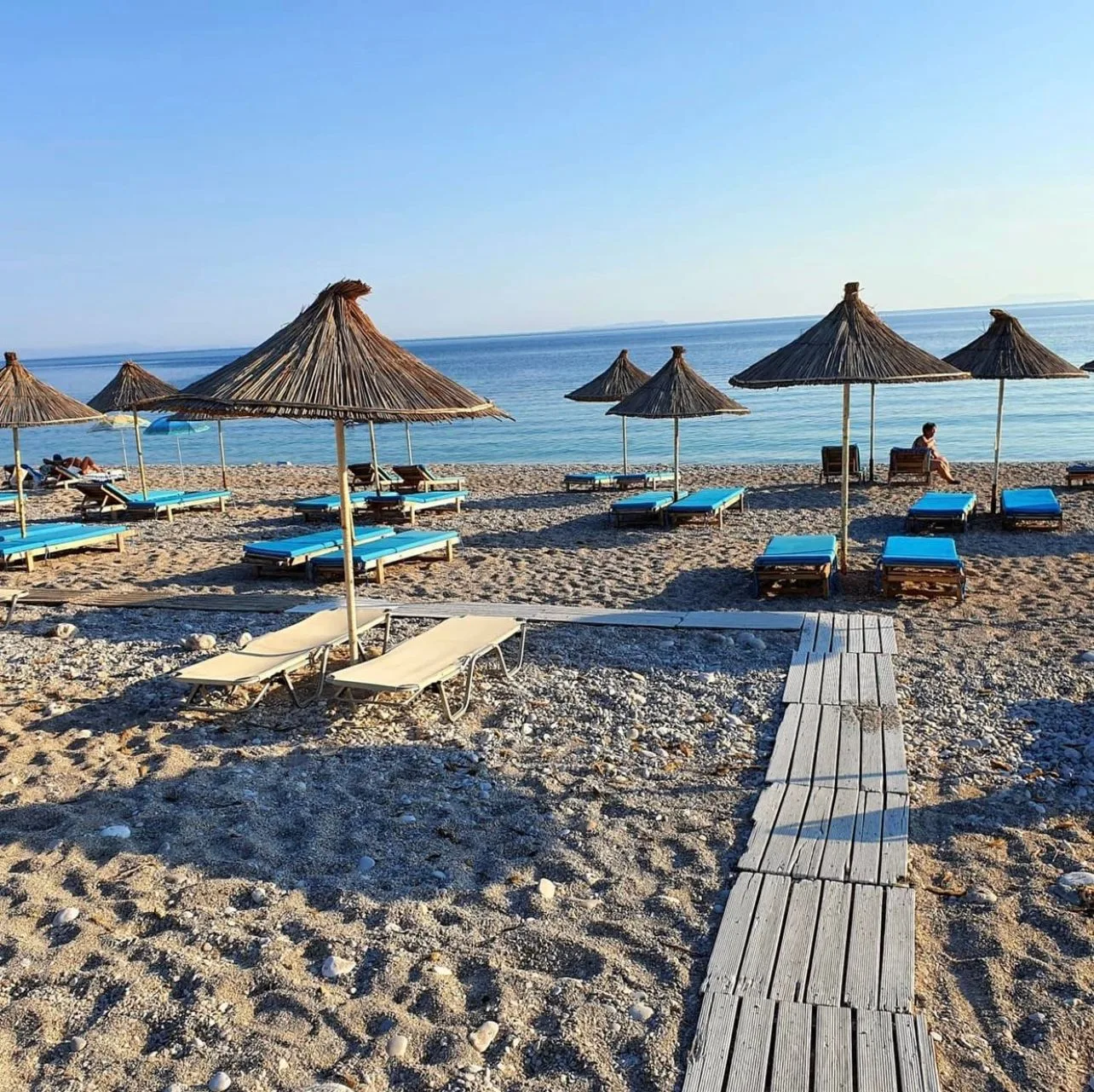 A plank boardwalk running across the shingle between straw umbrellas and blue-cushioned sunbeds to the water