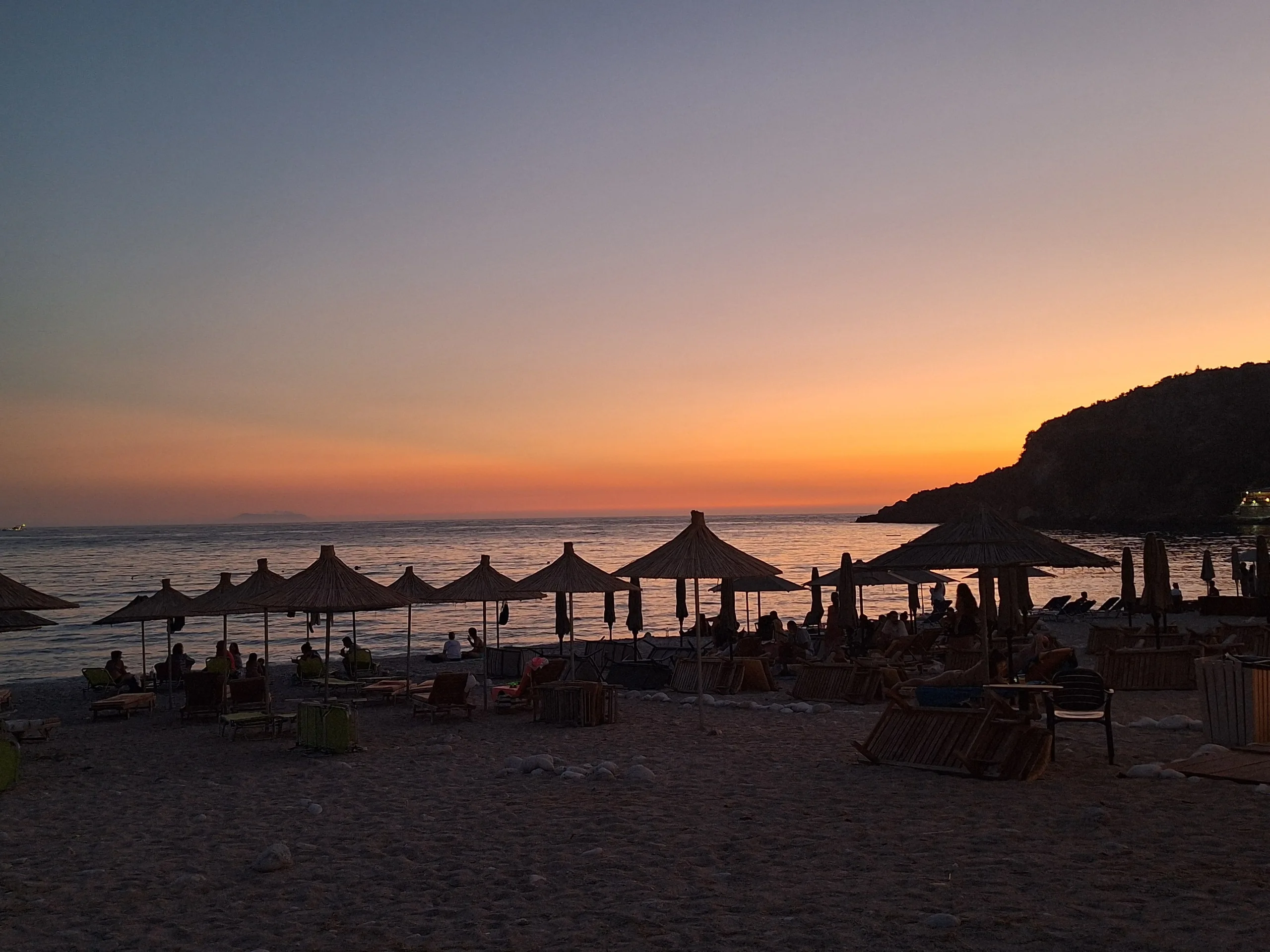 The same beach after sunset: straw umbrellas in silhouette against an orange horizon, the headland dark on the right