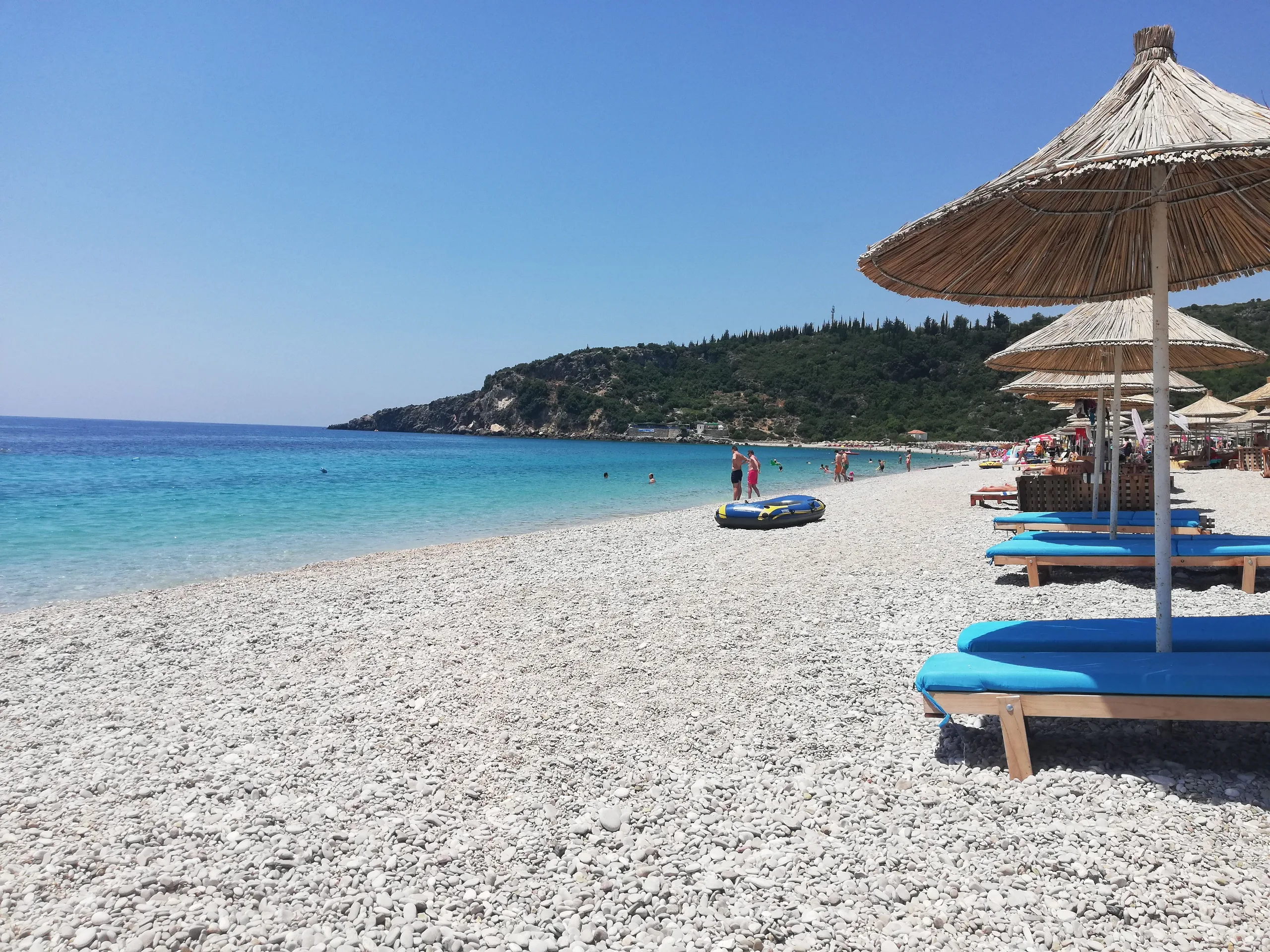 Livadh Beach at midday: pale shingle, straw umbrellas over blue-cushioned sunbeds, clear turquoise water and the wooded headland at the north end of the bay