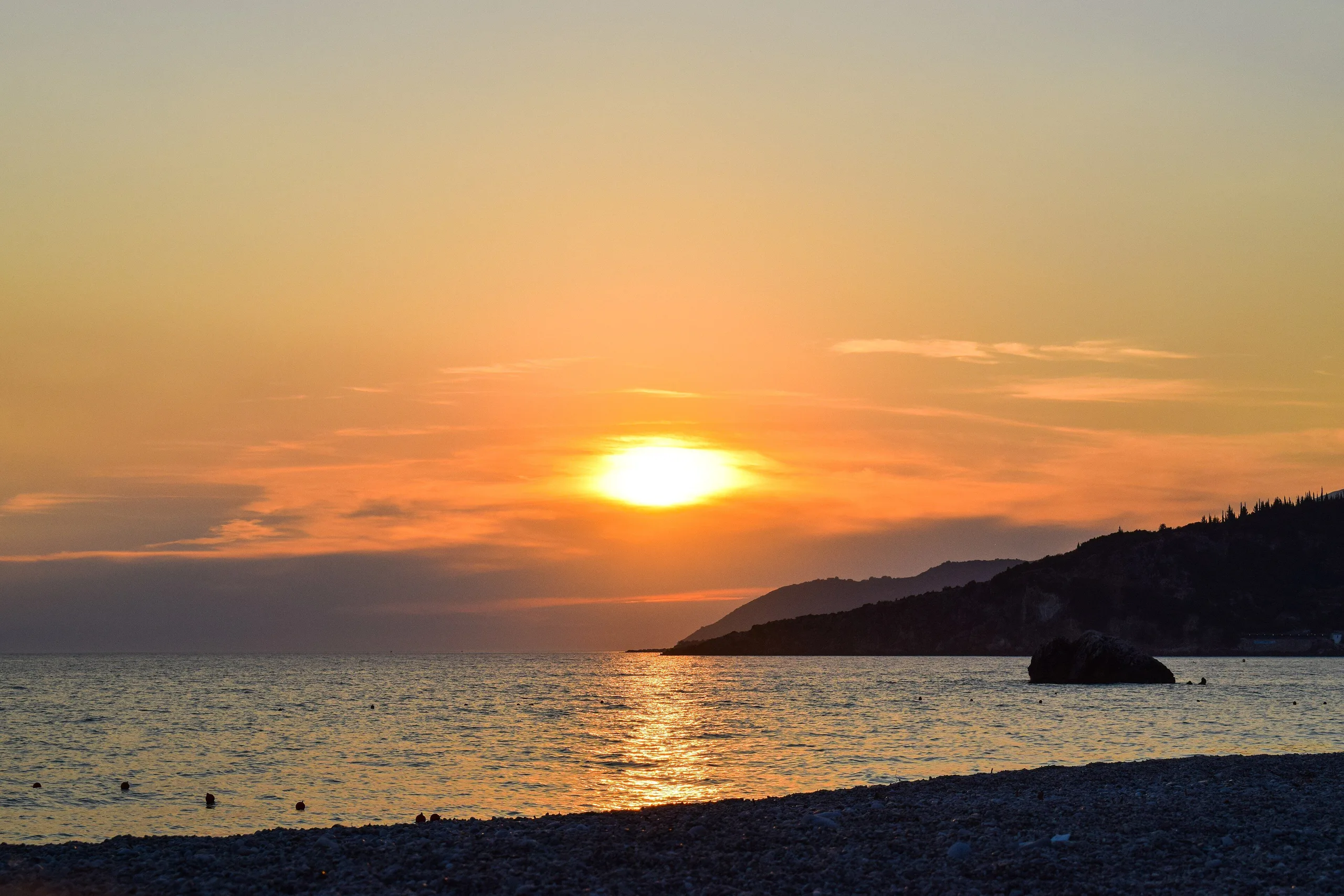 The sun setting into the sea off Livadh Beach, the headland and its offshore rock dark against the orange sky