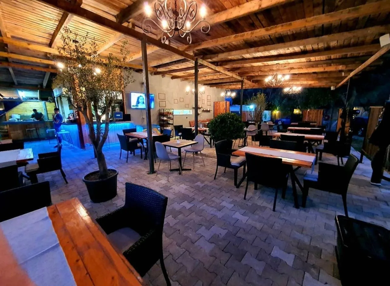 The camp's covered terrace at night: timber roof, wrought-iron chandeliers, a potted olive tree, tables set out on pavers and the bar lit blue at the back