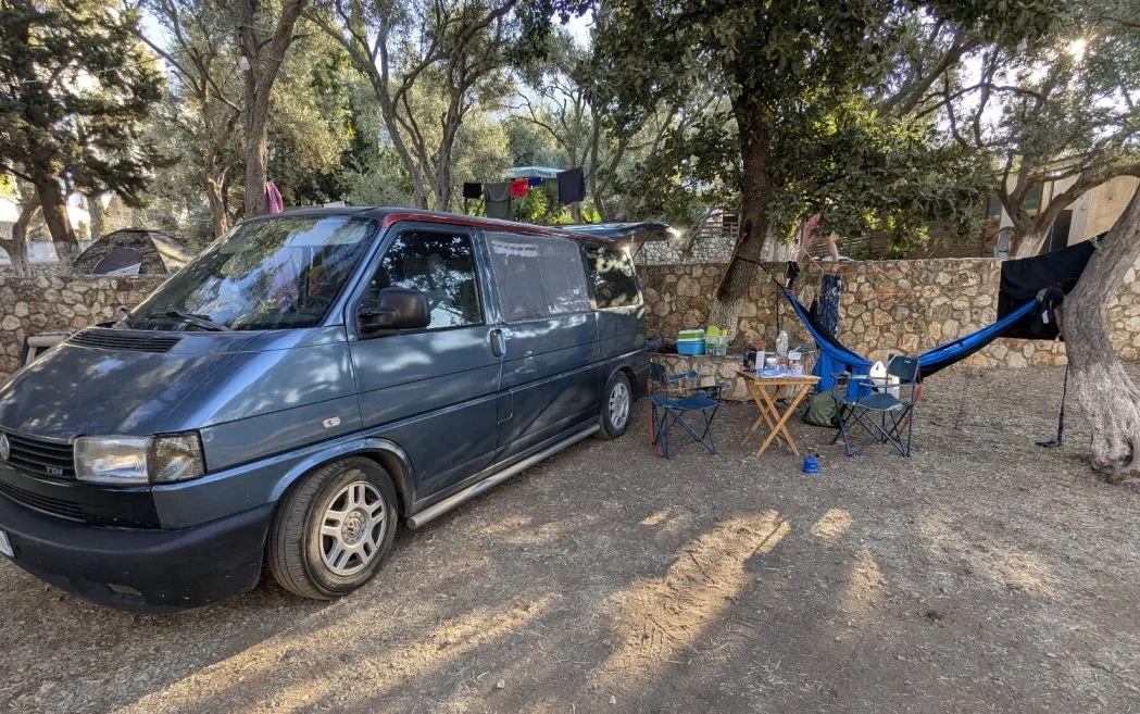 A grey van on a gravel pitch by a dry-stone wall, with a camping table, chairs and a hammock slung between two trees
