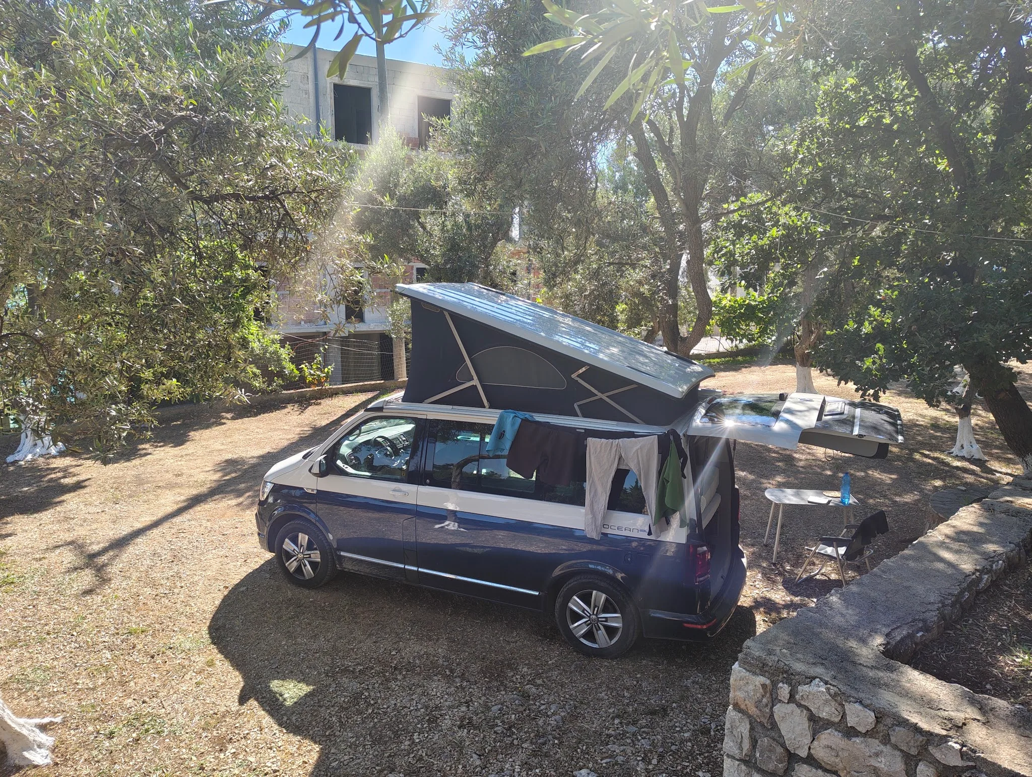 A pop-top campervan parked on dry ground in olive shade beside a low stone wall, a folding table set up next to it