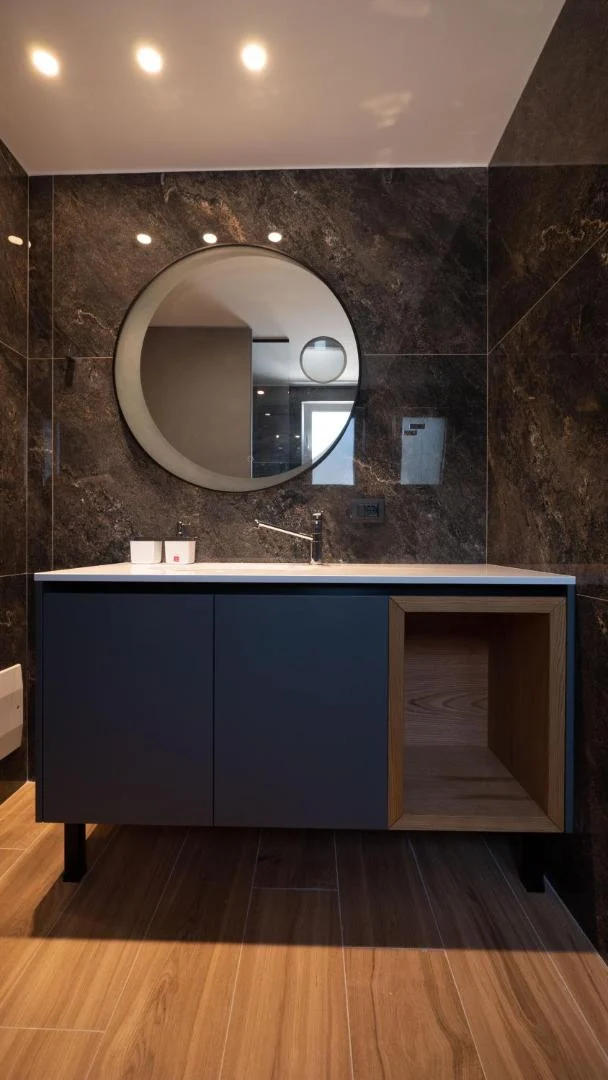 A bathroom lined in bronze-brown marble-effect tile with a round black-framed mirror, a navy vanity unit with an open oak cubby, and a wood-look plank floor